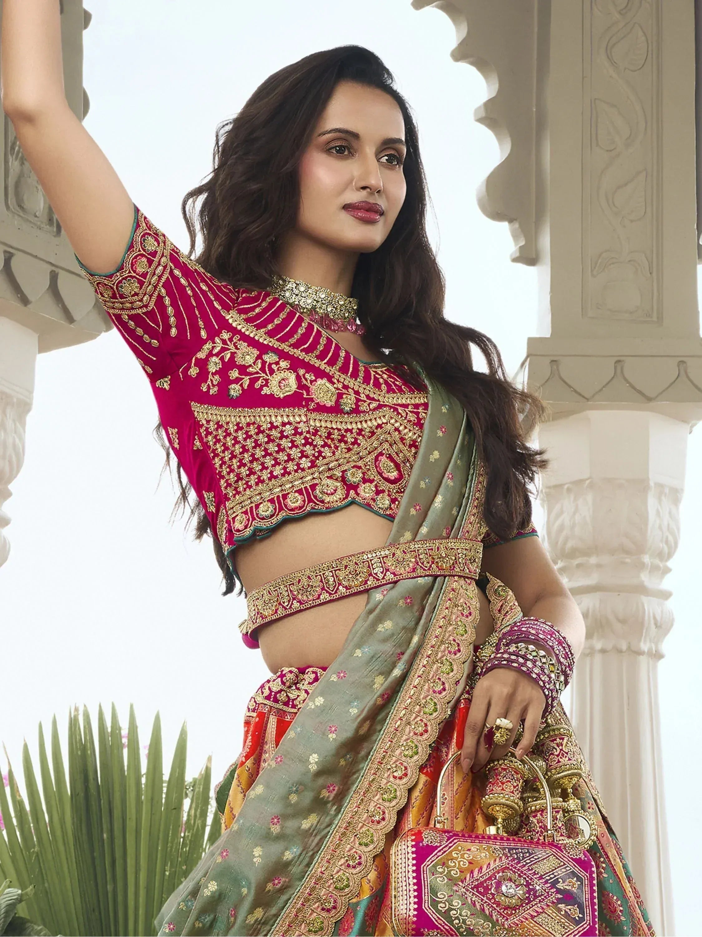 Woman in vibrant pink and green embroidered lehenga choli with gold detailing, holding matching handbag