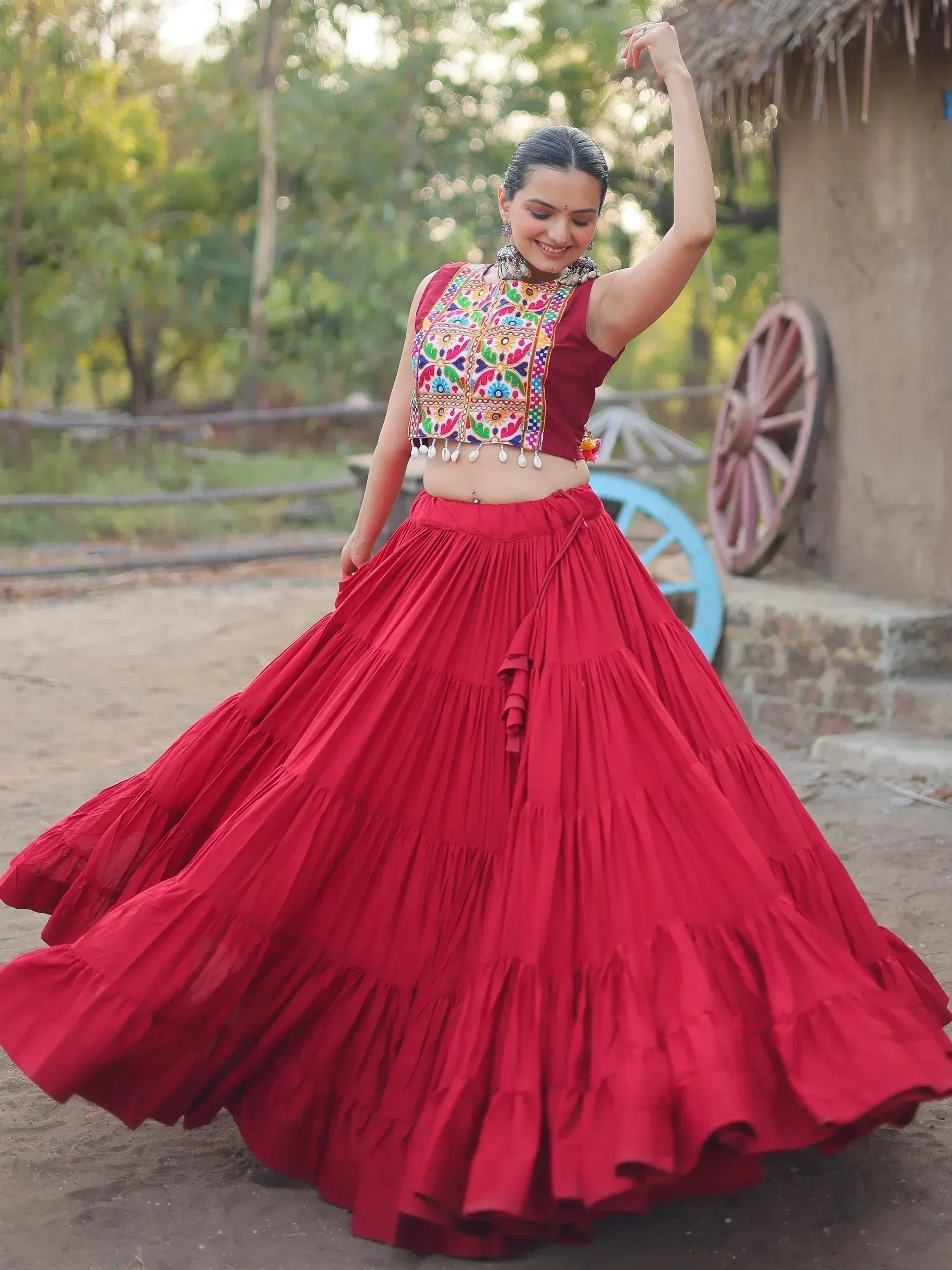 Woman in vibrant red ethnic lehenga with colorful embroidered blouse, dancing outdoors near rustic hut.
