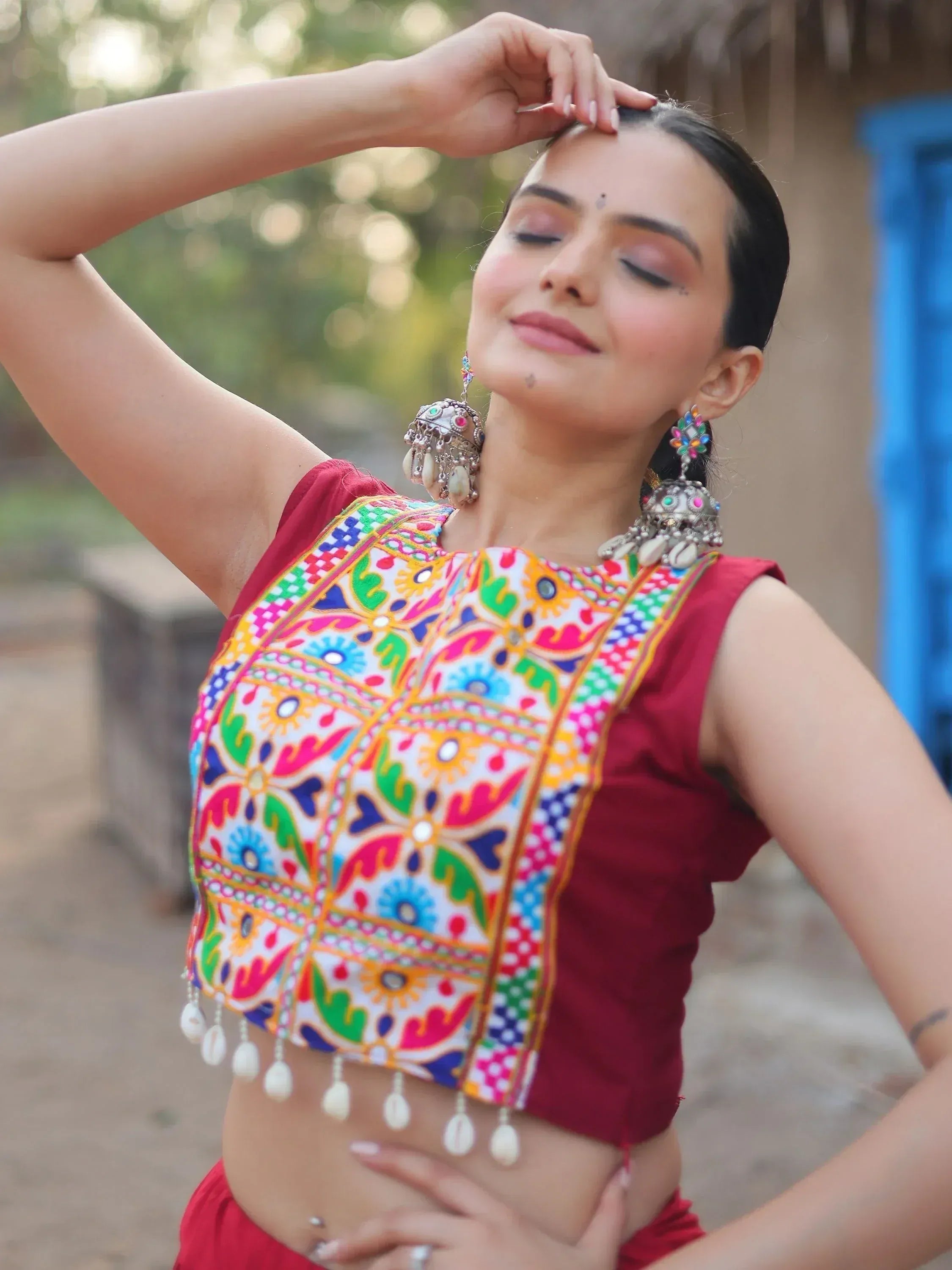 Woman in vibrant Indian ethnic crop top with mirror work and shell tassels, paired with traditional earrings.