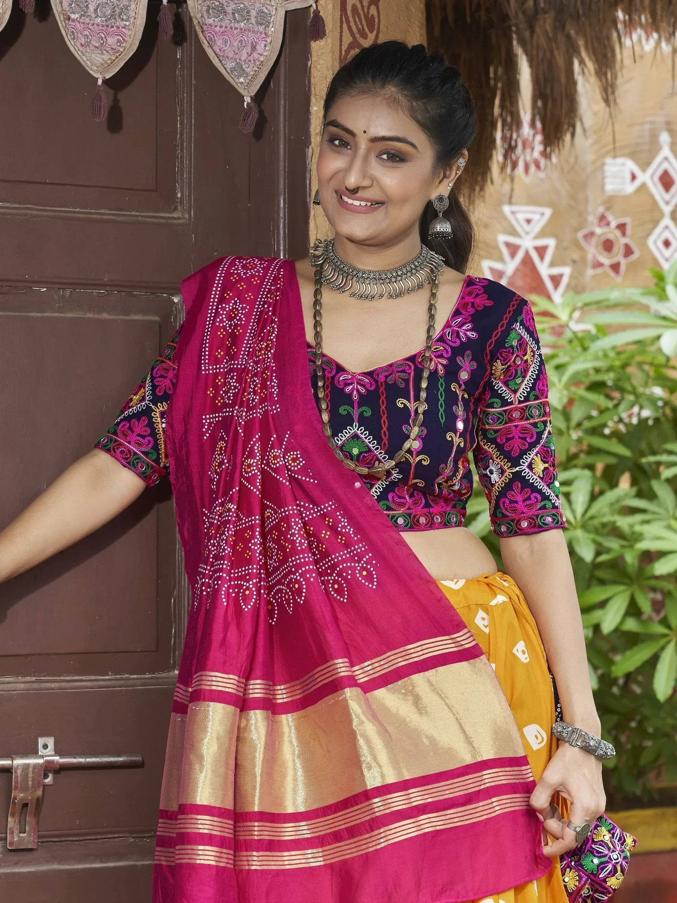 Woman in vibrant Indian ethnic wear with embroidered blouse and pink dupatta, traditional jewelry, rustic backdrop