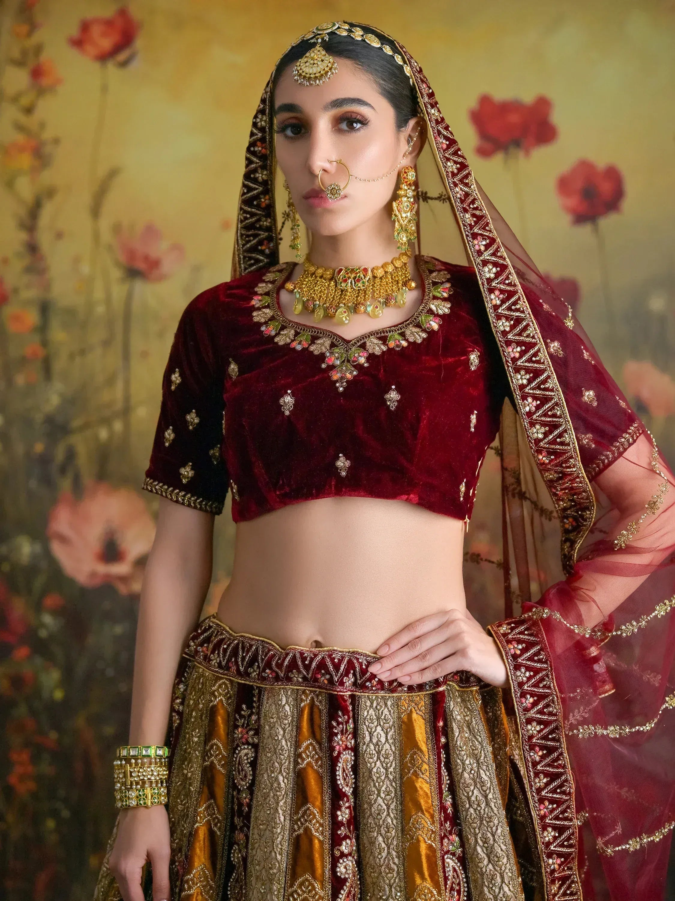 Maroon velvet lehenga choli with gold embroidery, traditional Indian bridal wear, heavy jewelry, floral backdrop