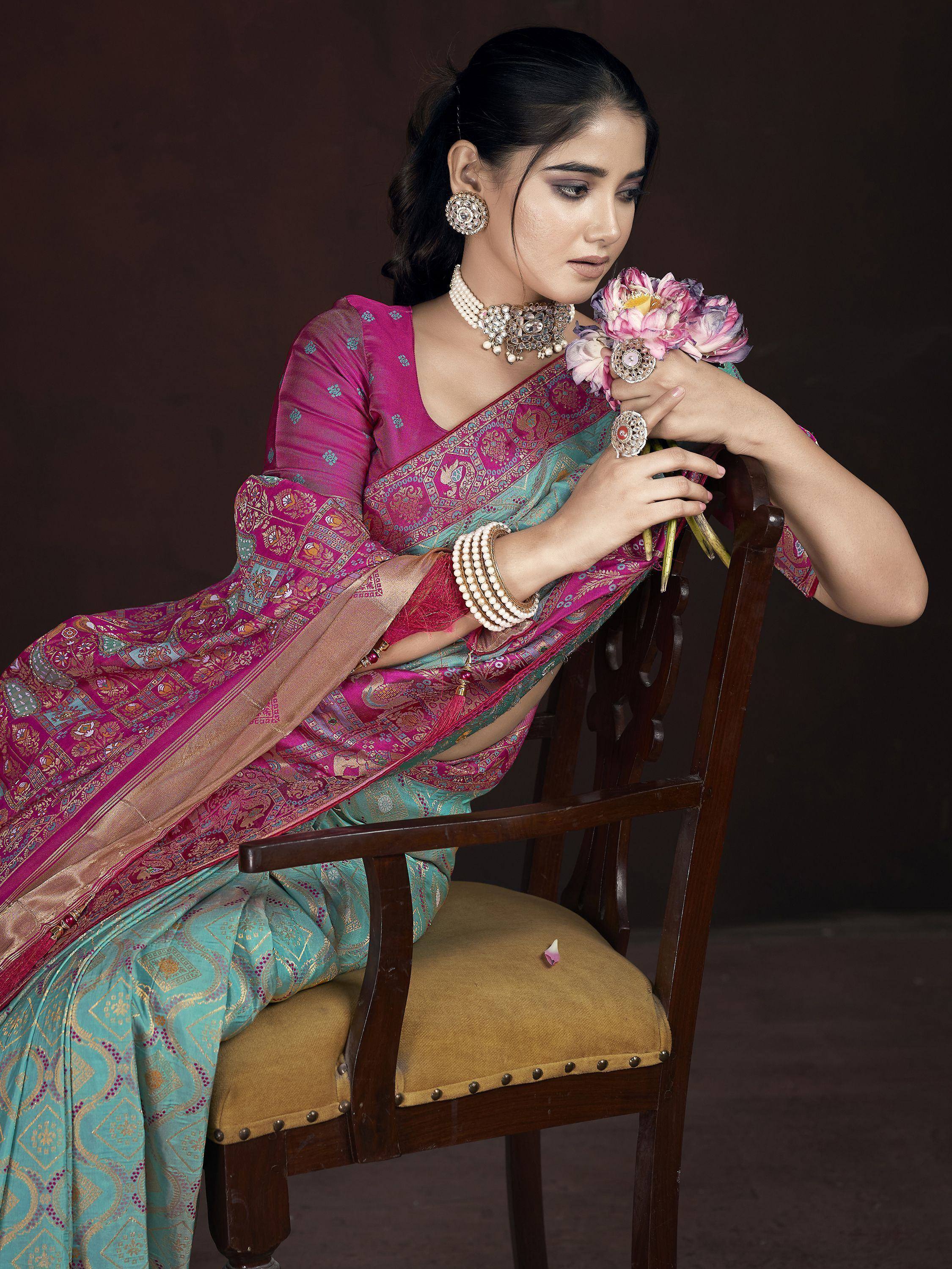 Woman in elegant pink and teal designer saree with gold embroidery, traditional jewelry, holding flowers
