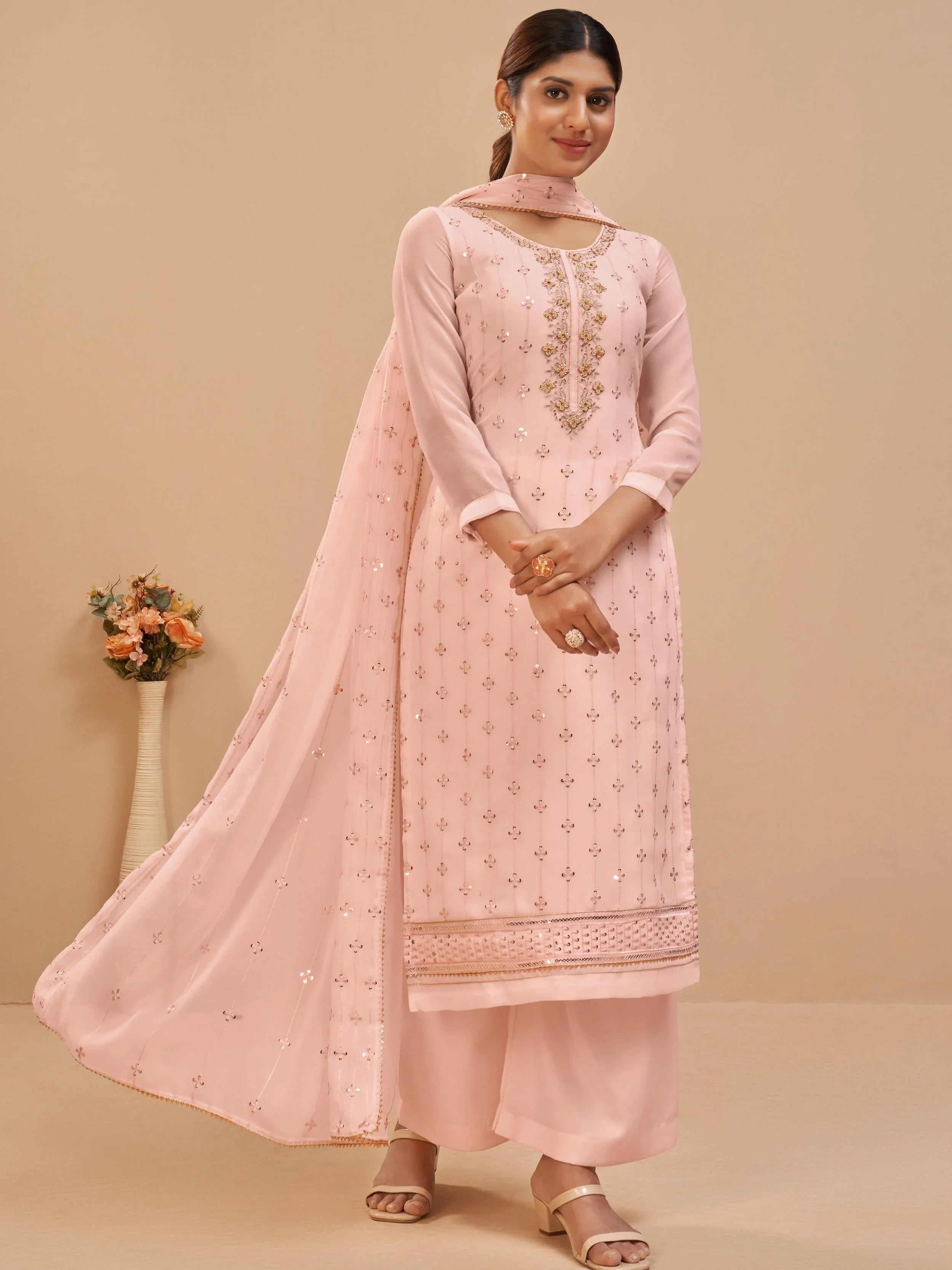 Woman in light pink embroidered salwar kameez with matching dupatta, standing indoors.
