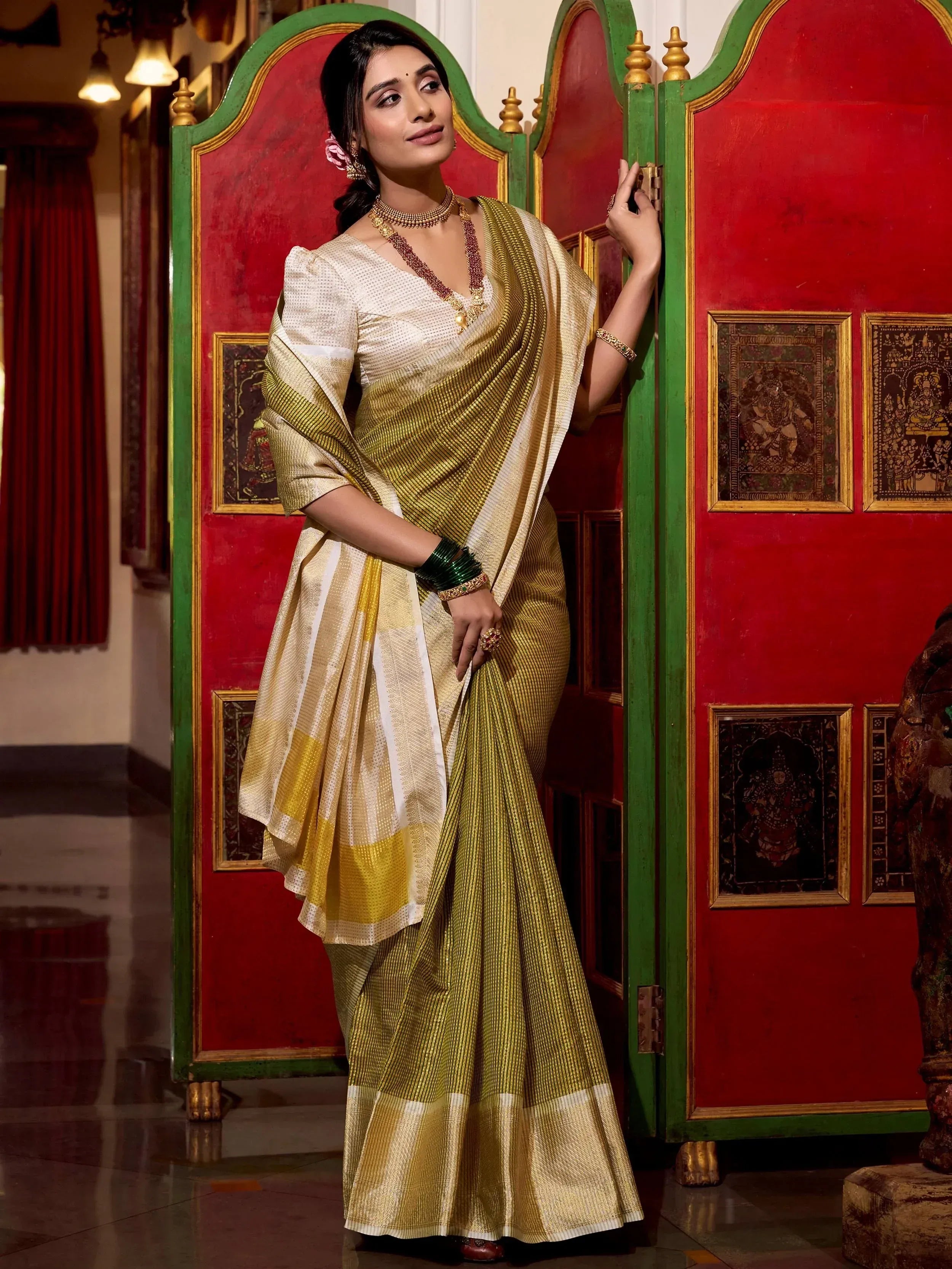 Elegant woman in golden striped saree with white blouse, traditional Indian ethnic wear, standing by ornate red and green door