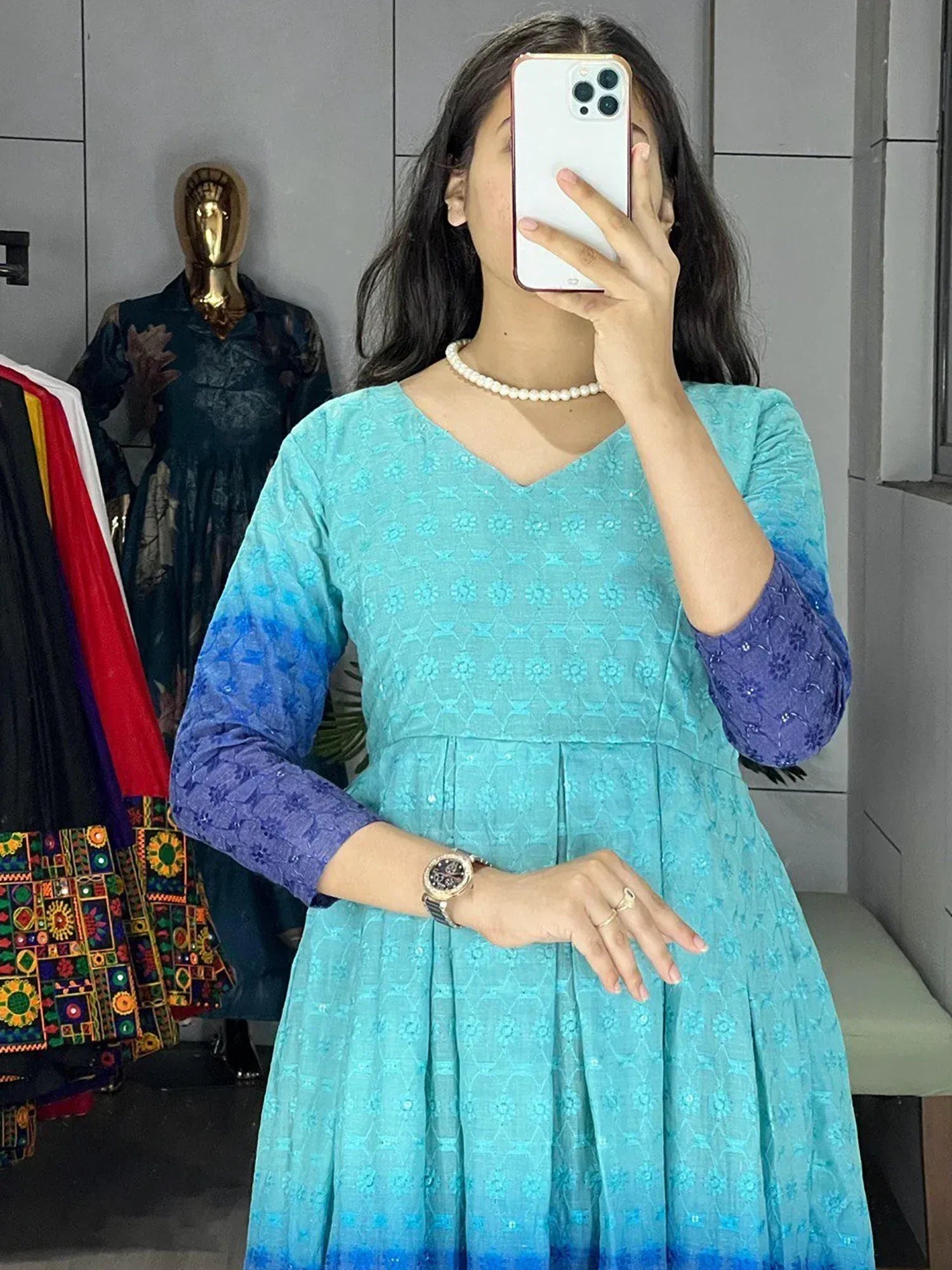Woman wearing blue ombre ethnic gown with floral print, pearl necklace, taking mirror selfie in boutique