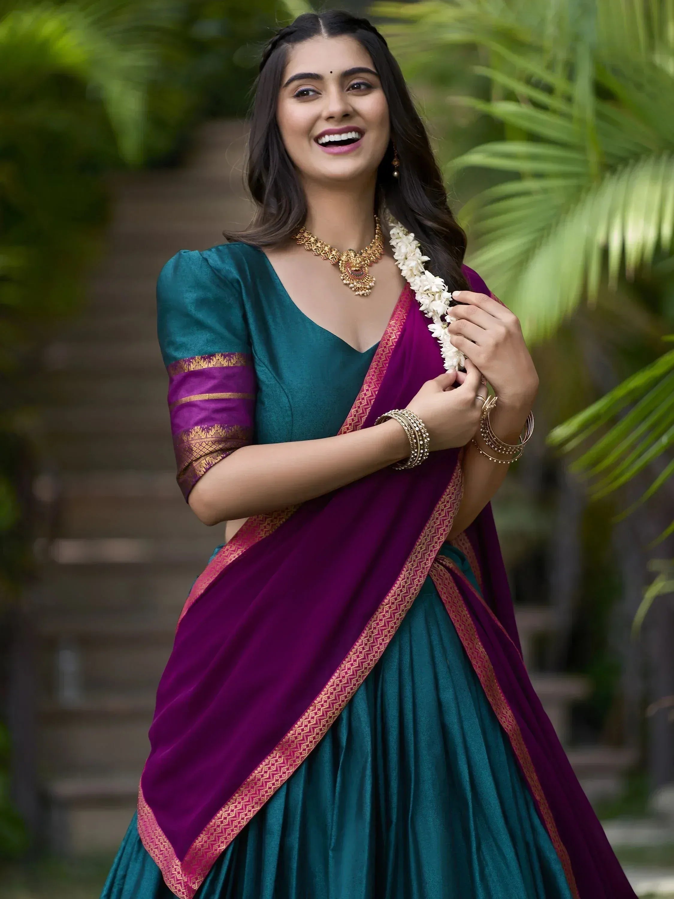 Woman in teal and purple Indian saree with gold border, floral garland, and traditional jewelry