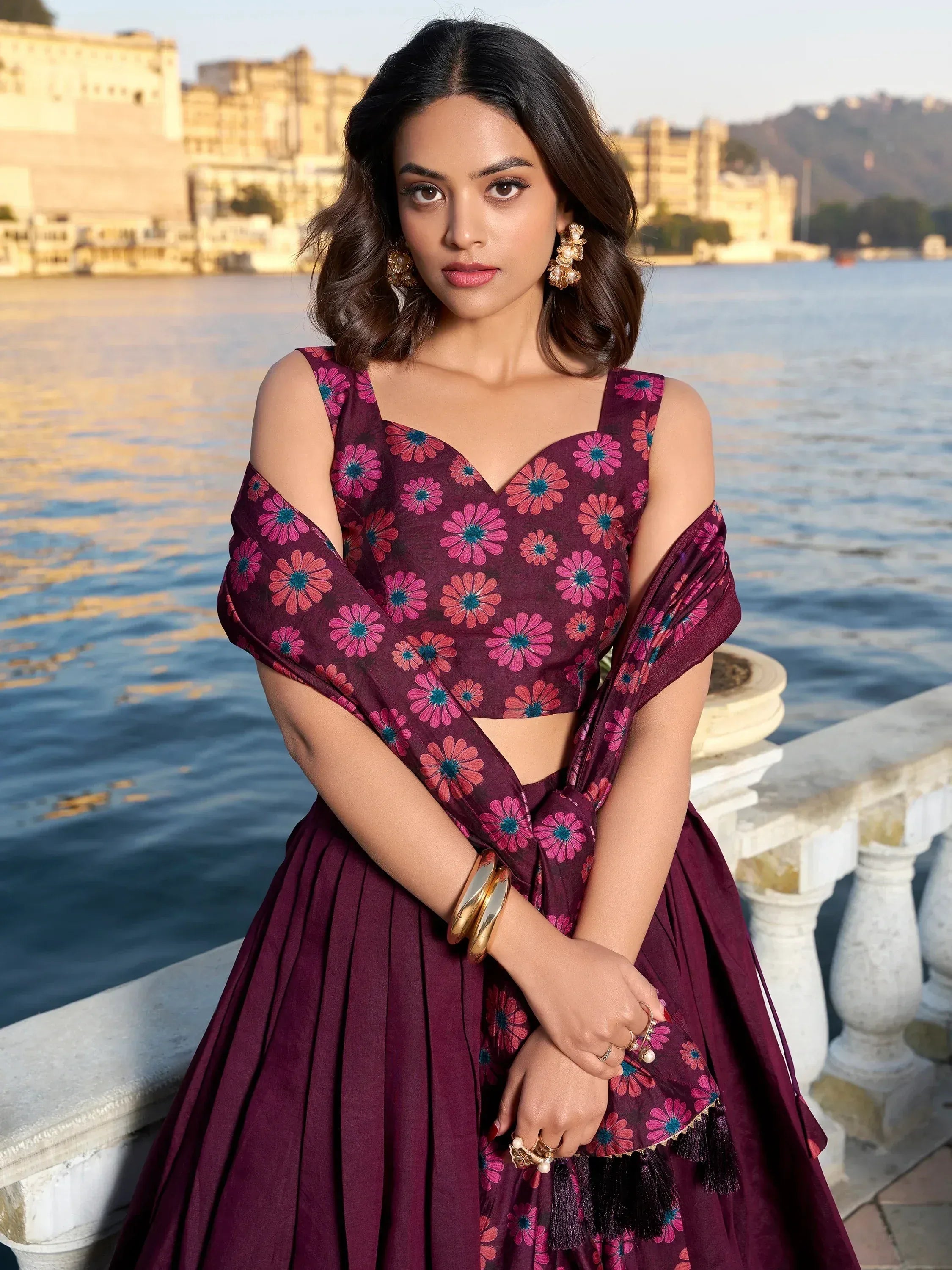 Woman in maroon floral lehenga choli with cold shoulder top, standing by lake with palace backdrop