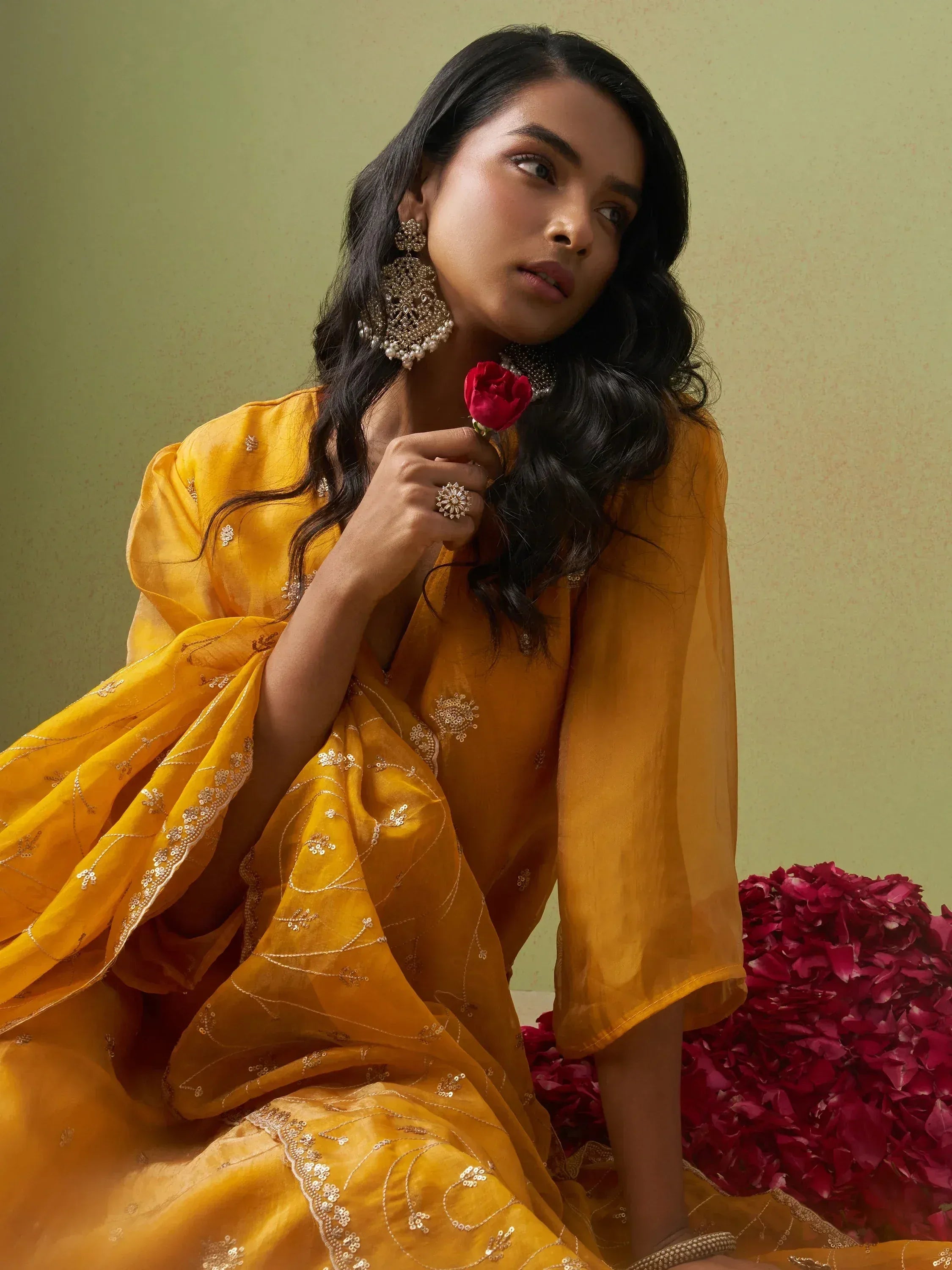 Woman in yellow embroidered ethnic gown with gold jewelry and red rose, Indian designer wear