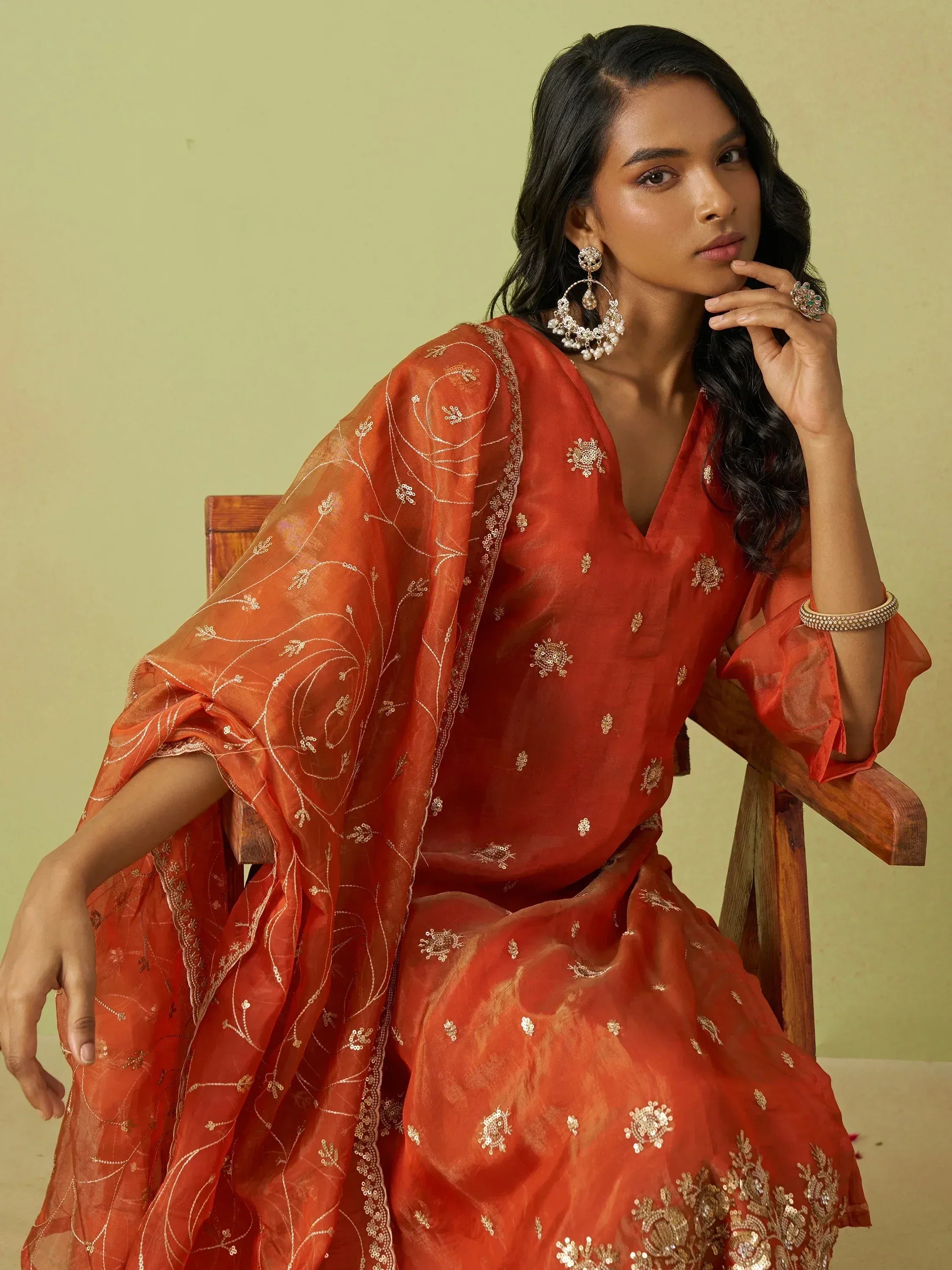 Woman in elegant orange ethnic gown with gold embroidery, styled with statement jewelry for festive occasions.