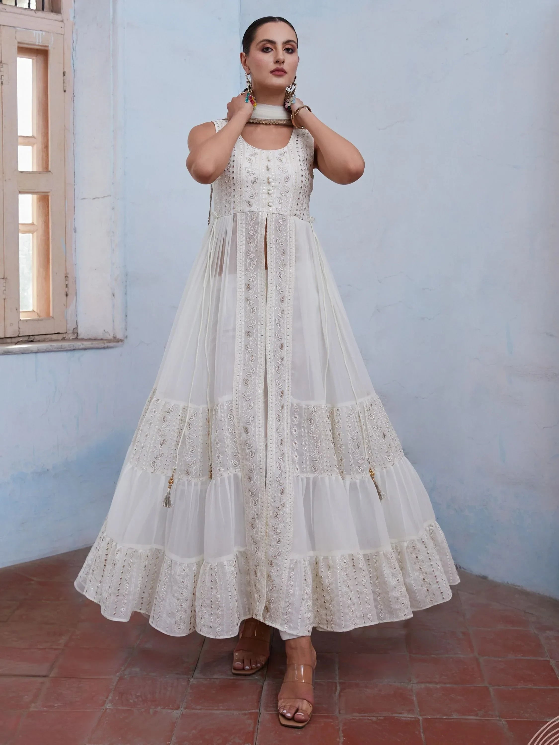 Woman wearing an embroidered white Anarkali gown with sheer panels, standing indoors.