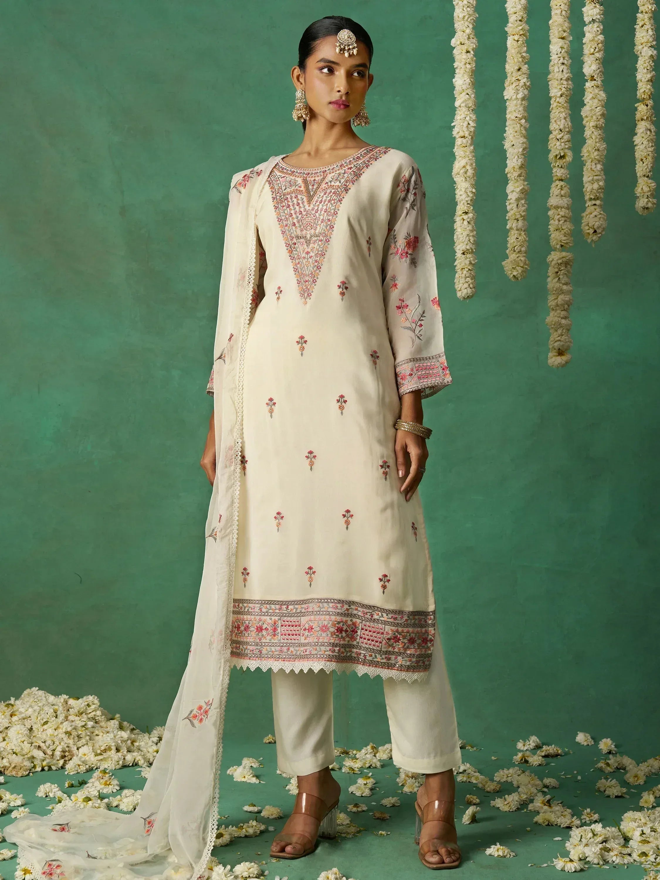 Cream embroidered salwar kameez with floral motifs, dupatta, and traditional jewelry against green backdrop with flowers
