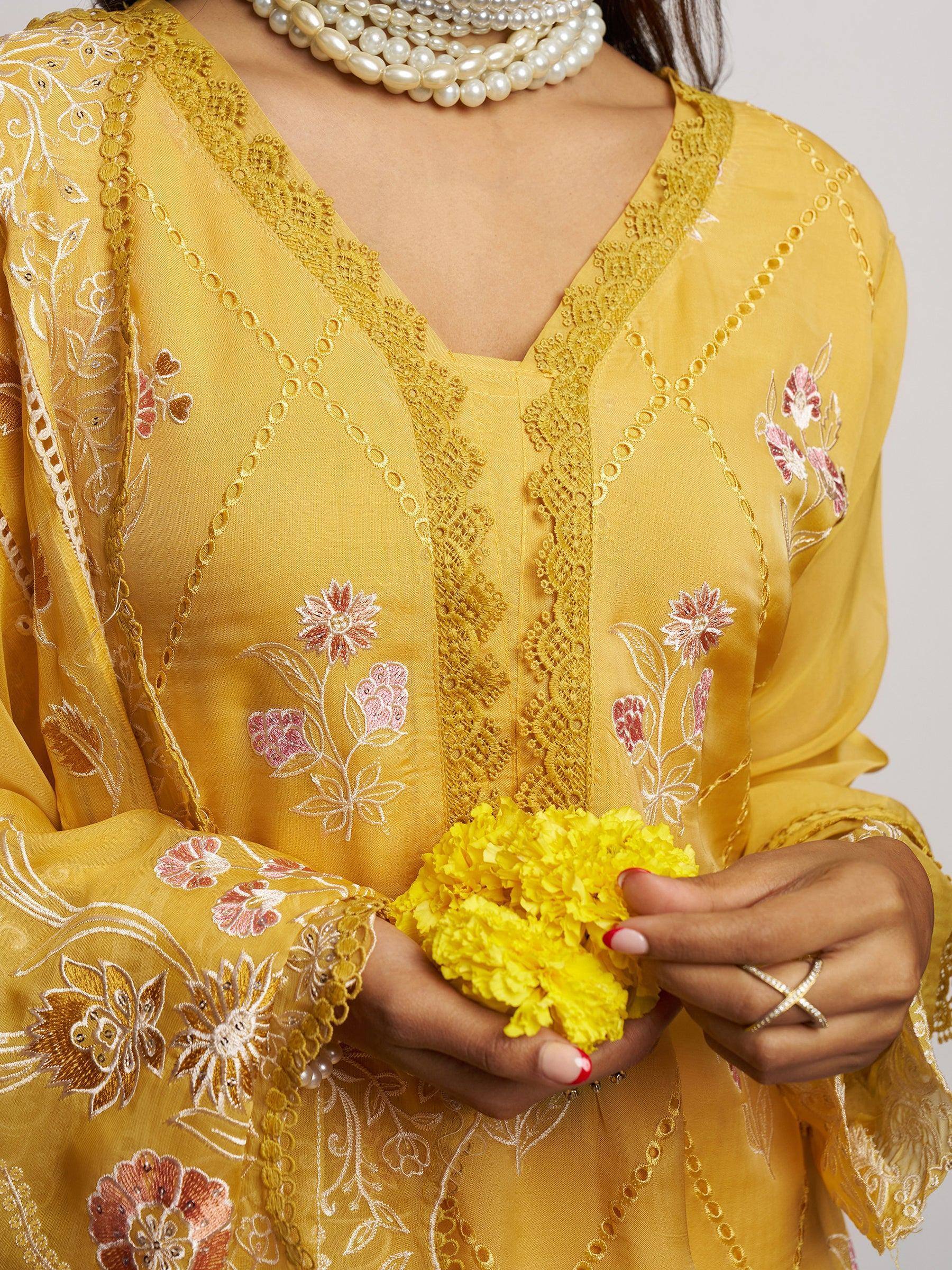 Yellow embroidered ethnic gown with floral details, lace trim, and pearl necklace, holding marigold flowers