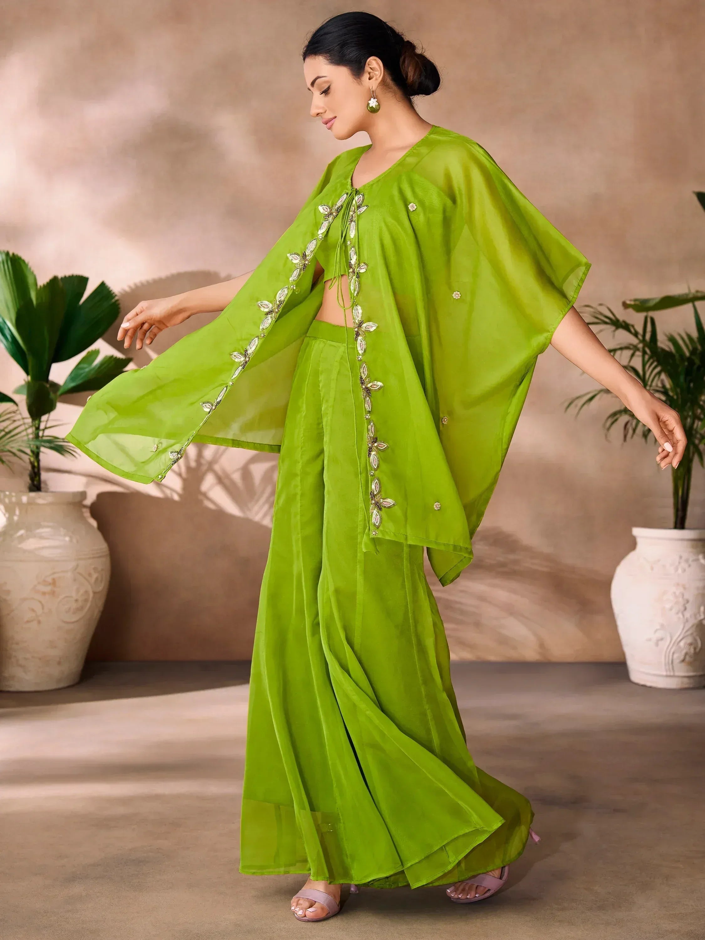 Woman in lime green ethnic gown with embroidered cape, flared pants, indoor setting, plants