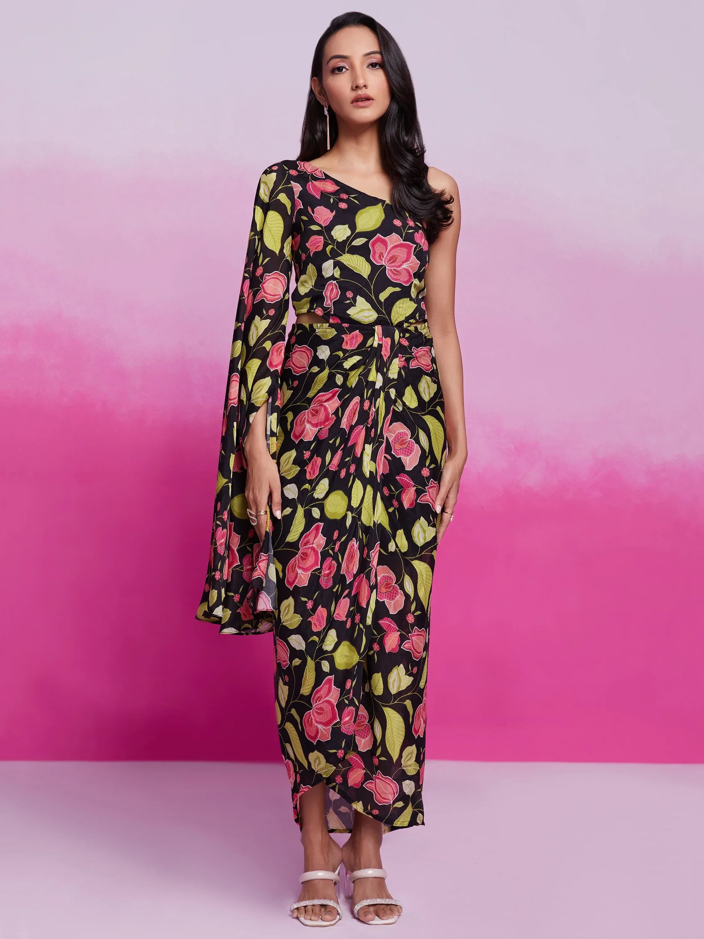Woman in one-shoulder floral print saree dress with pink and green patterns, standing indoors