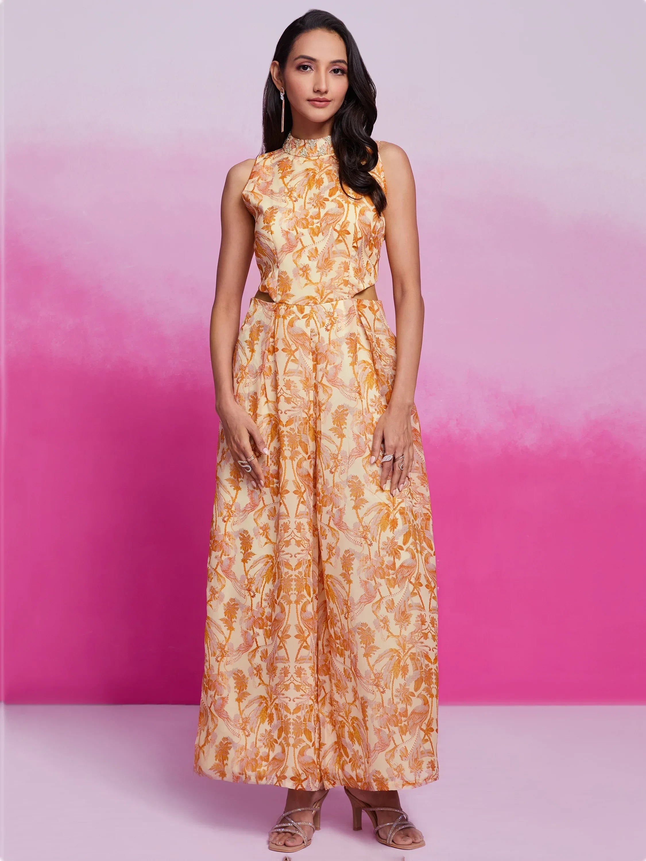Woman in a sleeveless yellow floral ethnic gown with high neck, standing against pink backdrop
