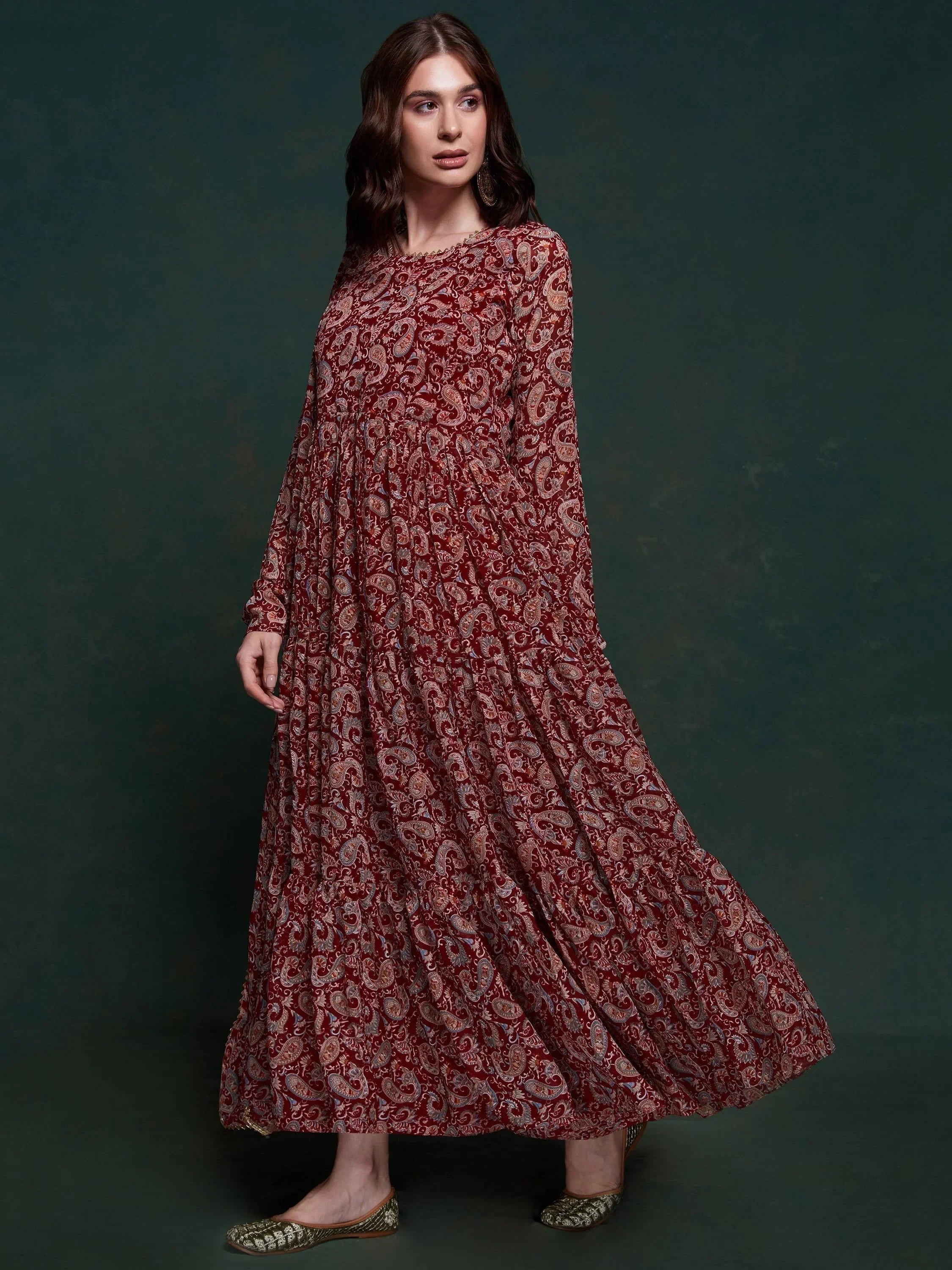 Woman wearing maroon paisley print ethnic gown with long sleeves, standing against dark background