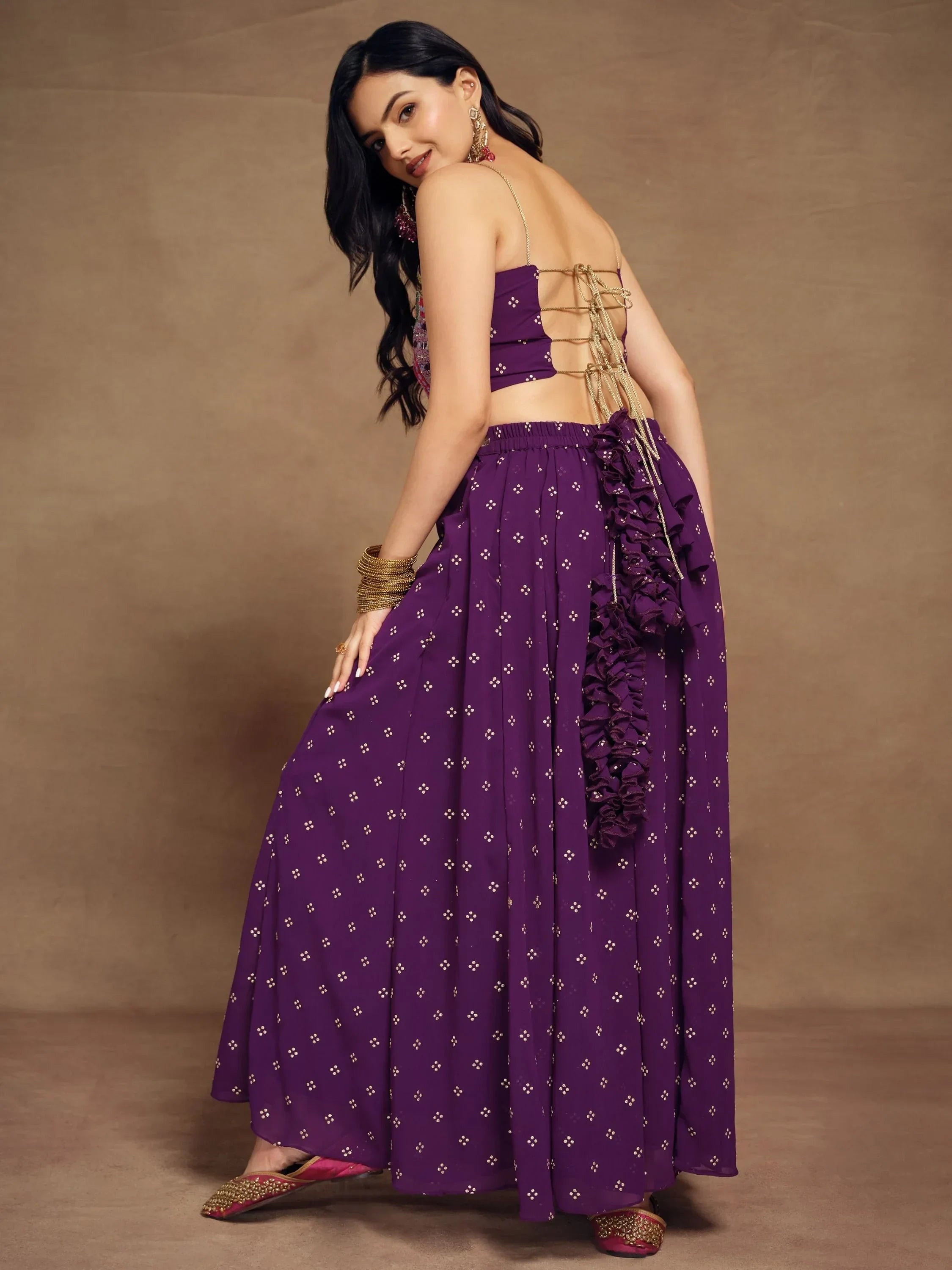 Woman in a purple backless lehenga choli with gold detailing, ethnic jewelry, and jutti shoes