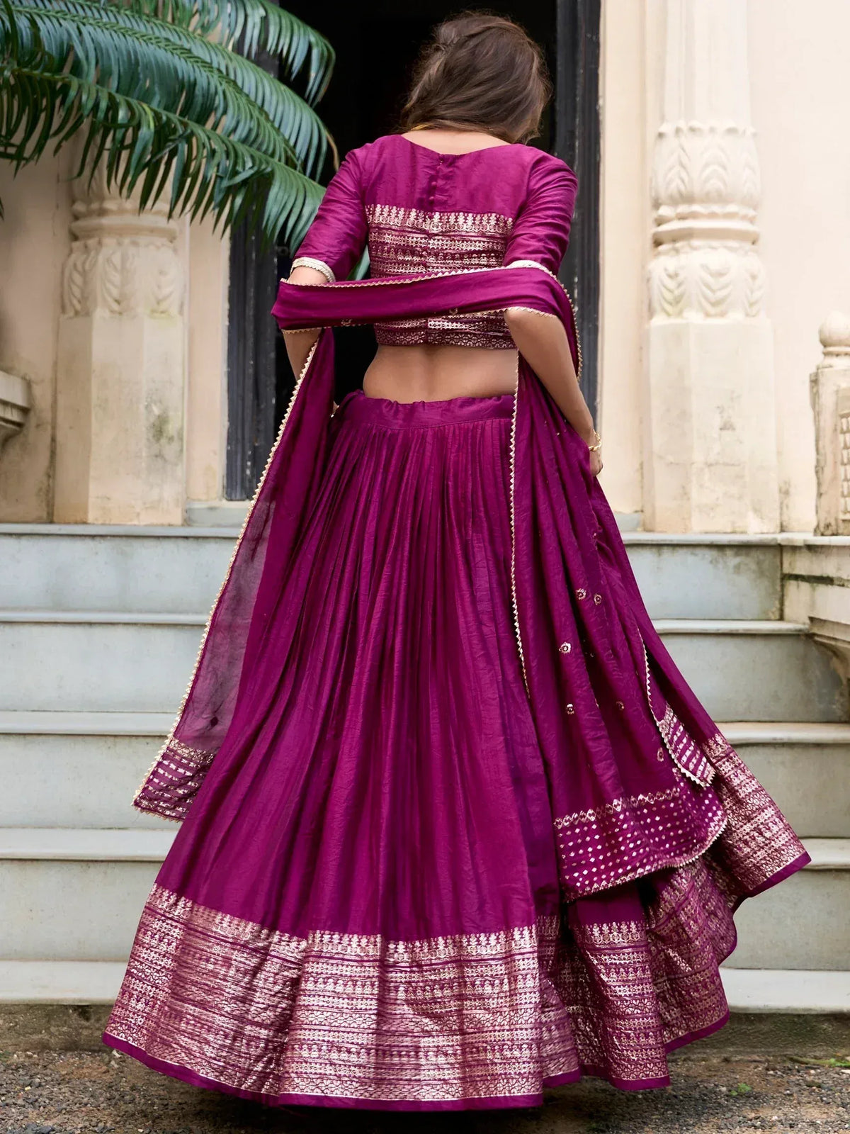 Purple designer lehenga choli with gold embroidery, back view on steps near palm tree