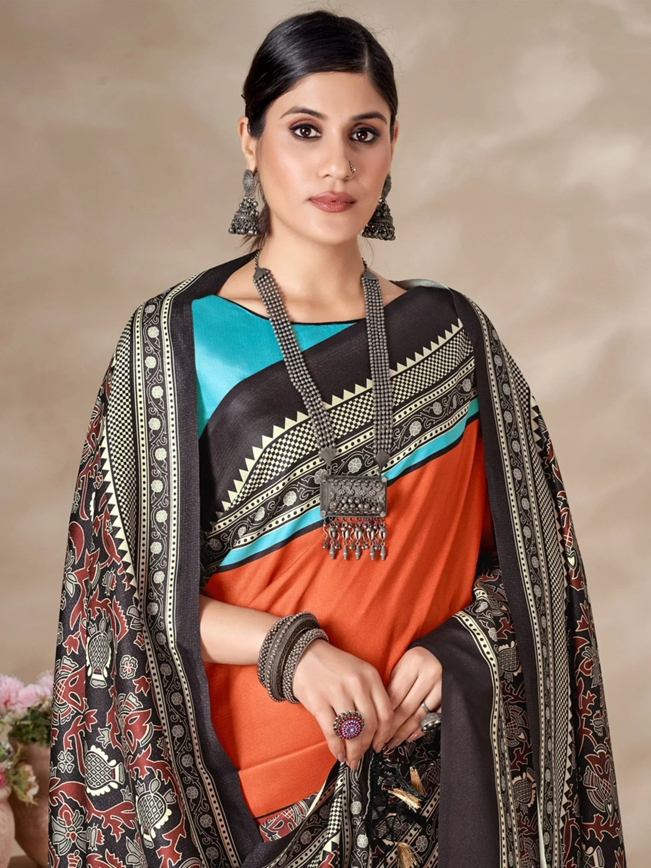 Woman in vibrant orange and teal saree with black printed border, ethnic jewelry, and traditional Indian attire