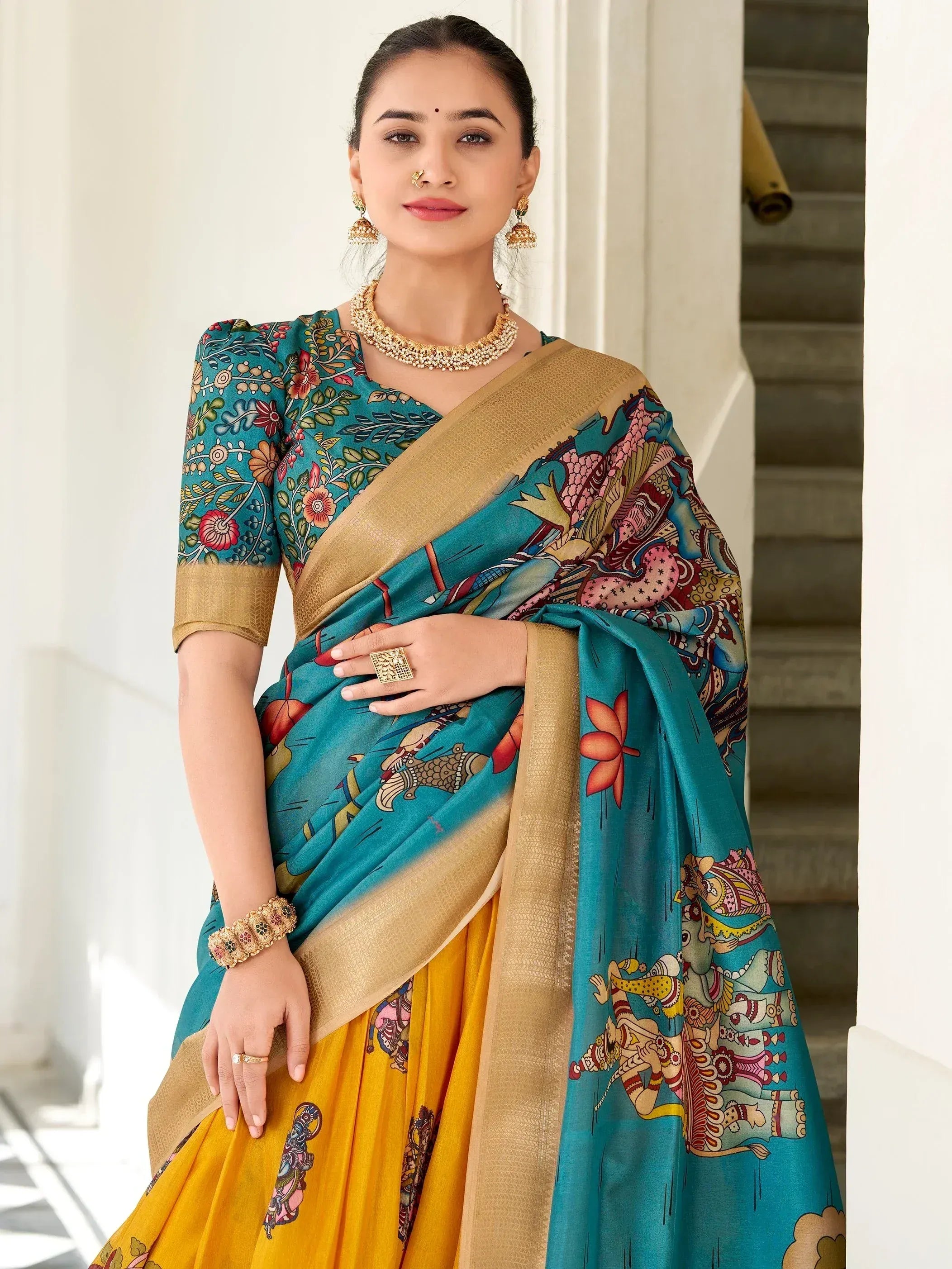 Teal and yellow printed saree with gold border, floral and mythological motifs, paired with traditional jewelry