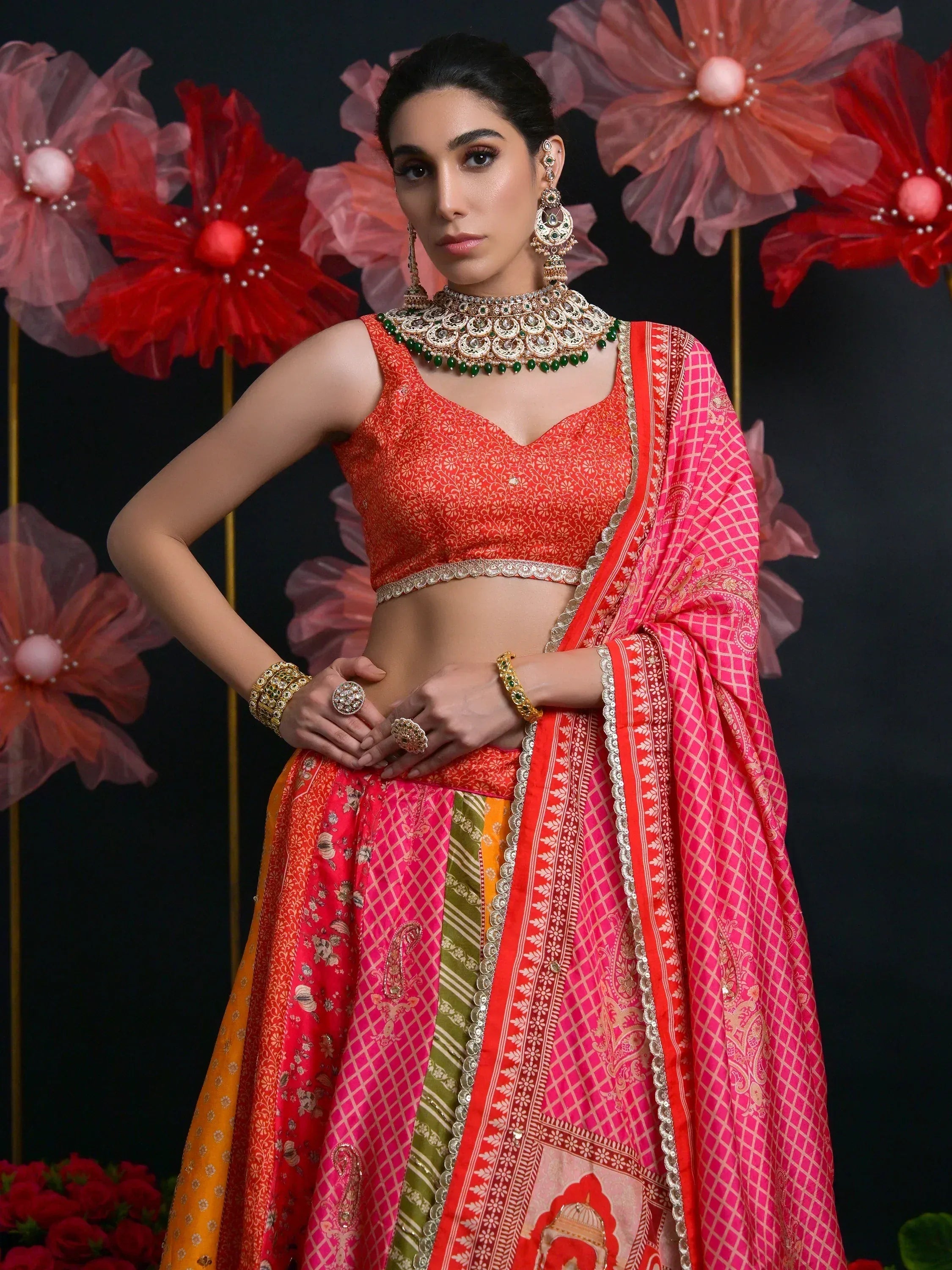 Vibrant orange and pink designer lehenga choli with intricate patterns, paired with traditional jewelry and floral backdrop.