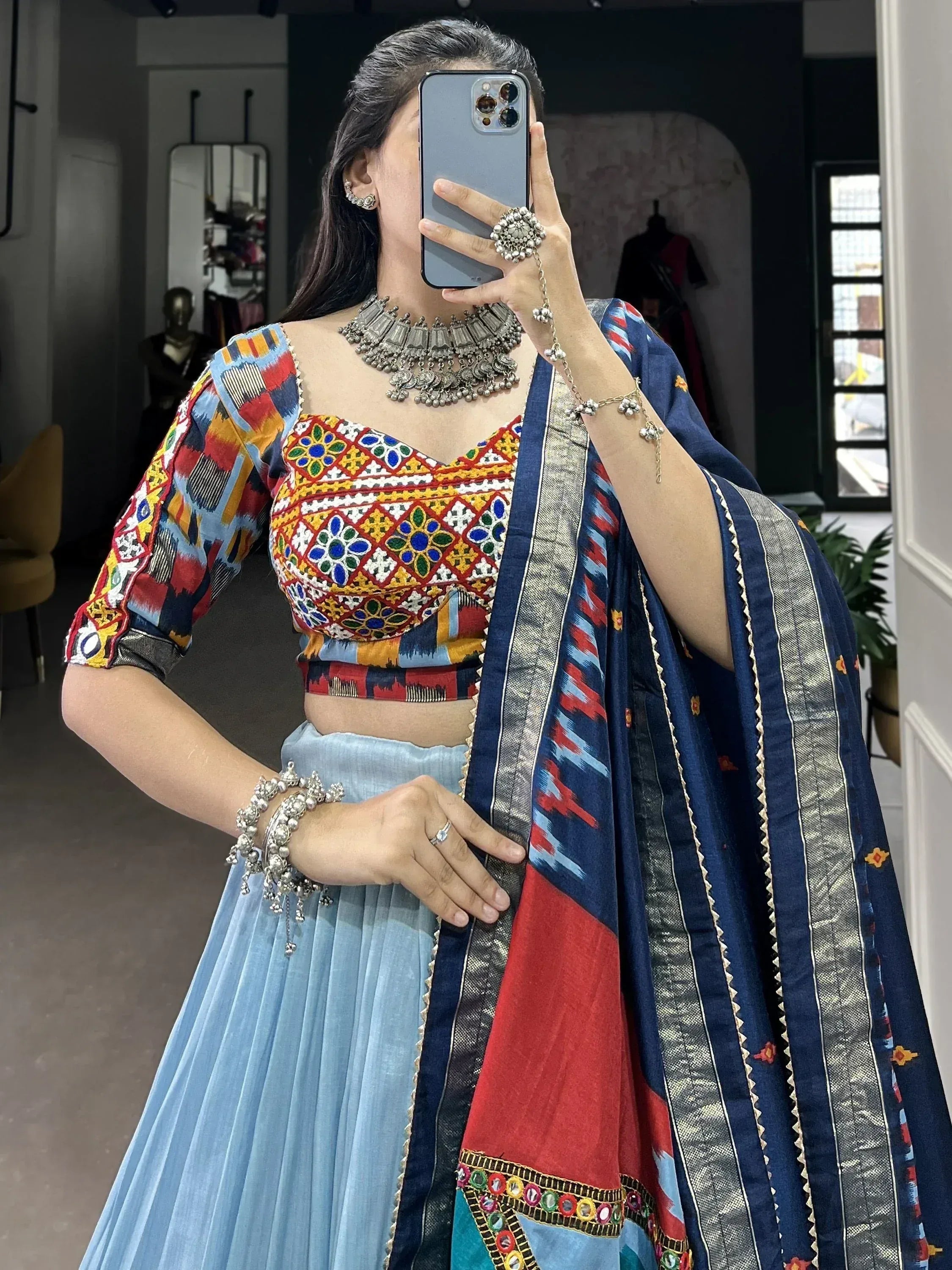 Woman in colorful embroidered lehenga choli with mirror work, paired with blue pleated skirt and dupatta, styled with silver jewelry.