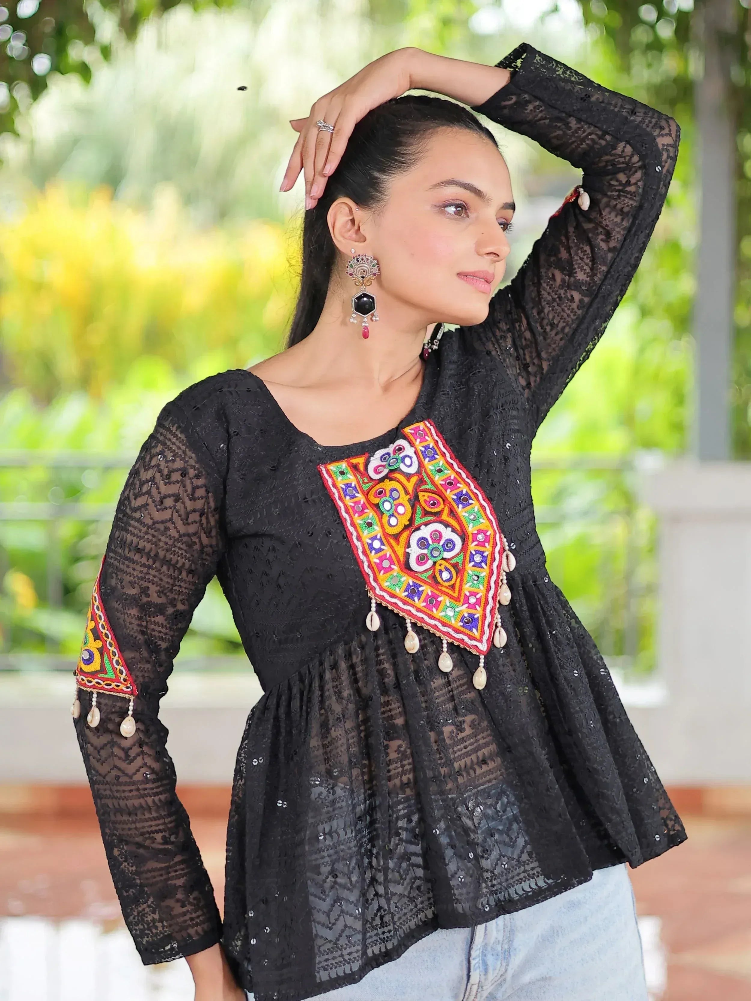 Black lace ethnic top with colorful mirror embroidery and shell tassels, paired with jeans for casual wear.
