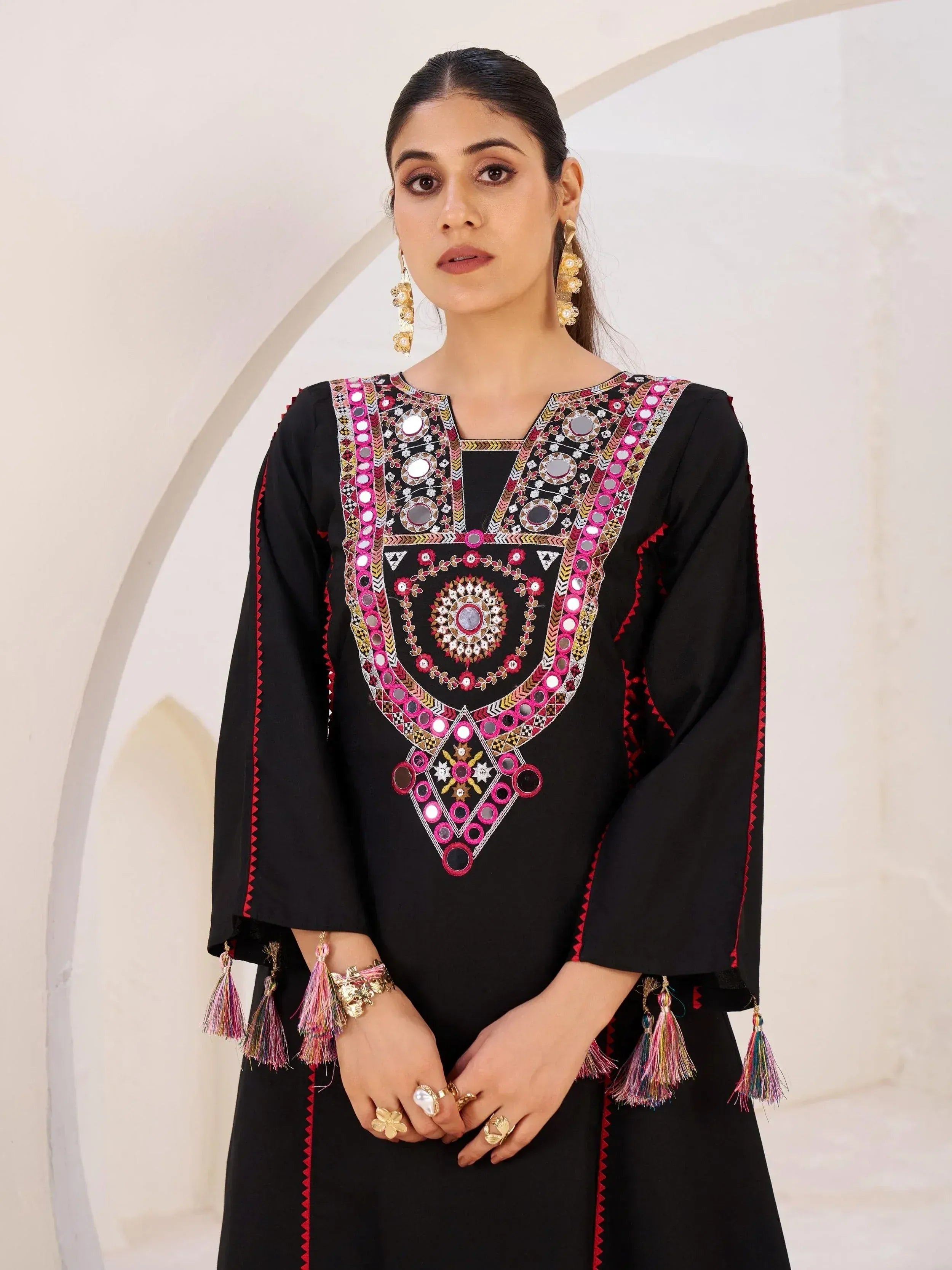Black ethnic kurti with mirror work, pink embroidery, and tassels, styled with gold jewelry