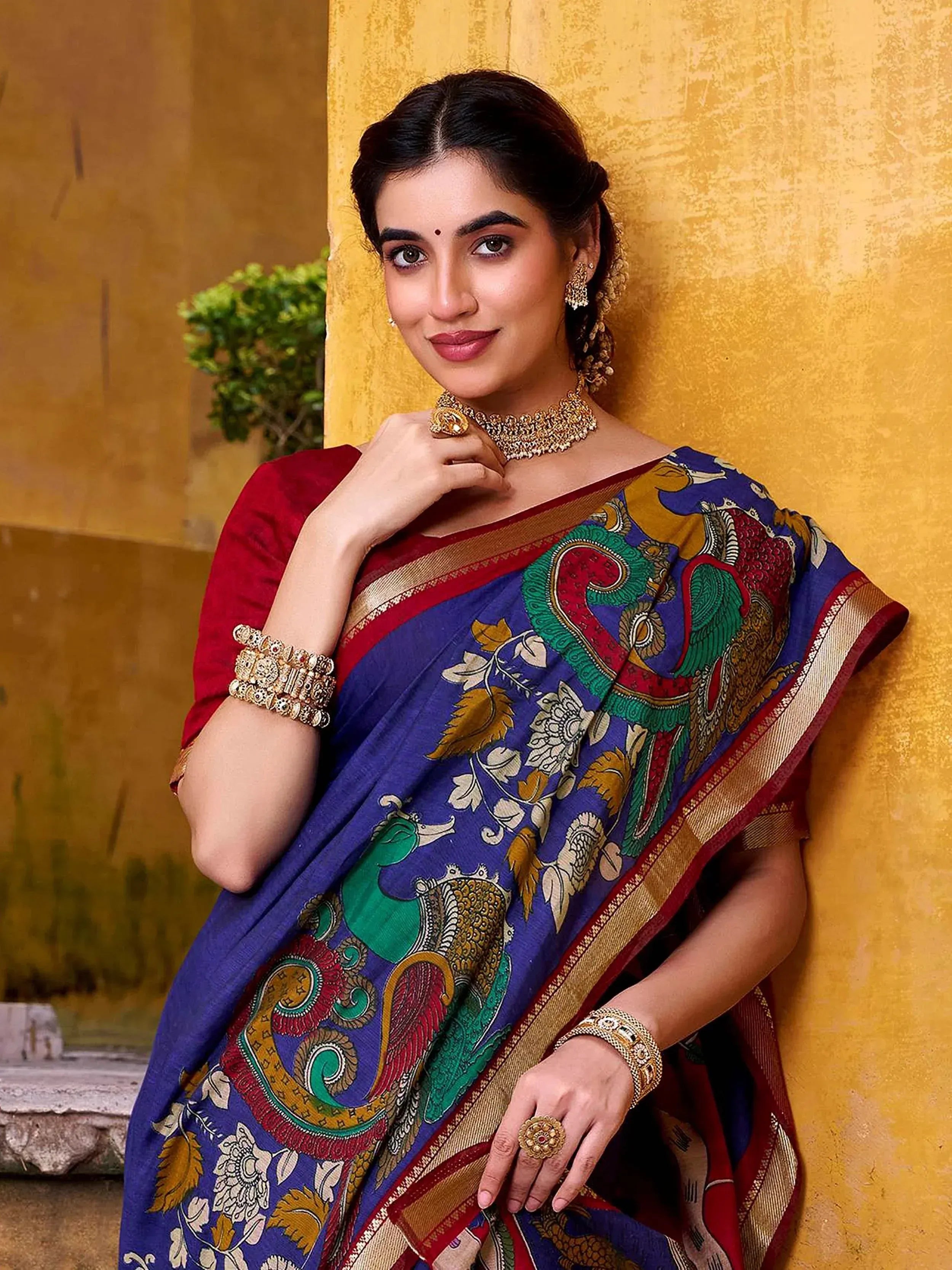 Woman in blue printed saree with peacock design, red blouse, gold jewelry, against yellow wall