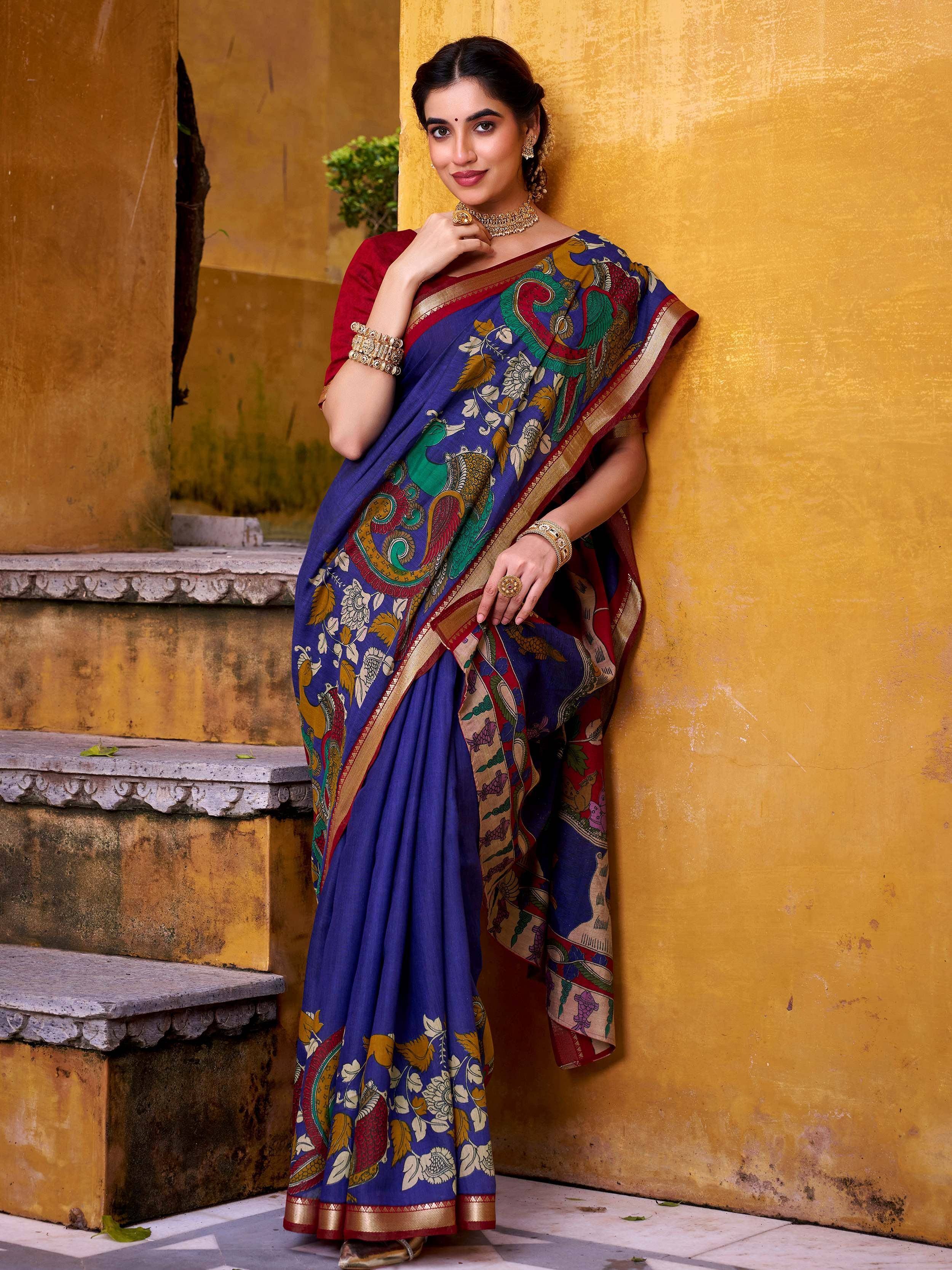 Woman in blue printed saree with peacock design, red blouse, gold jewelry, against yellow wall