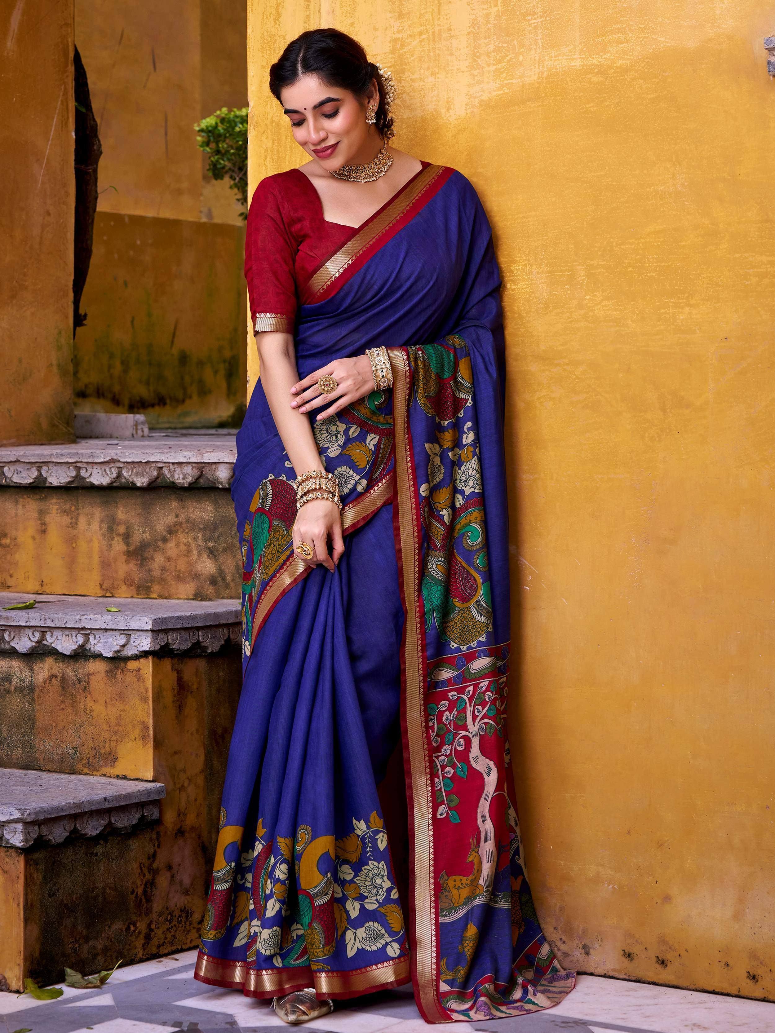 Woman in blue printed saree with red blouse, gold jewelry, against yellow wall