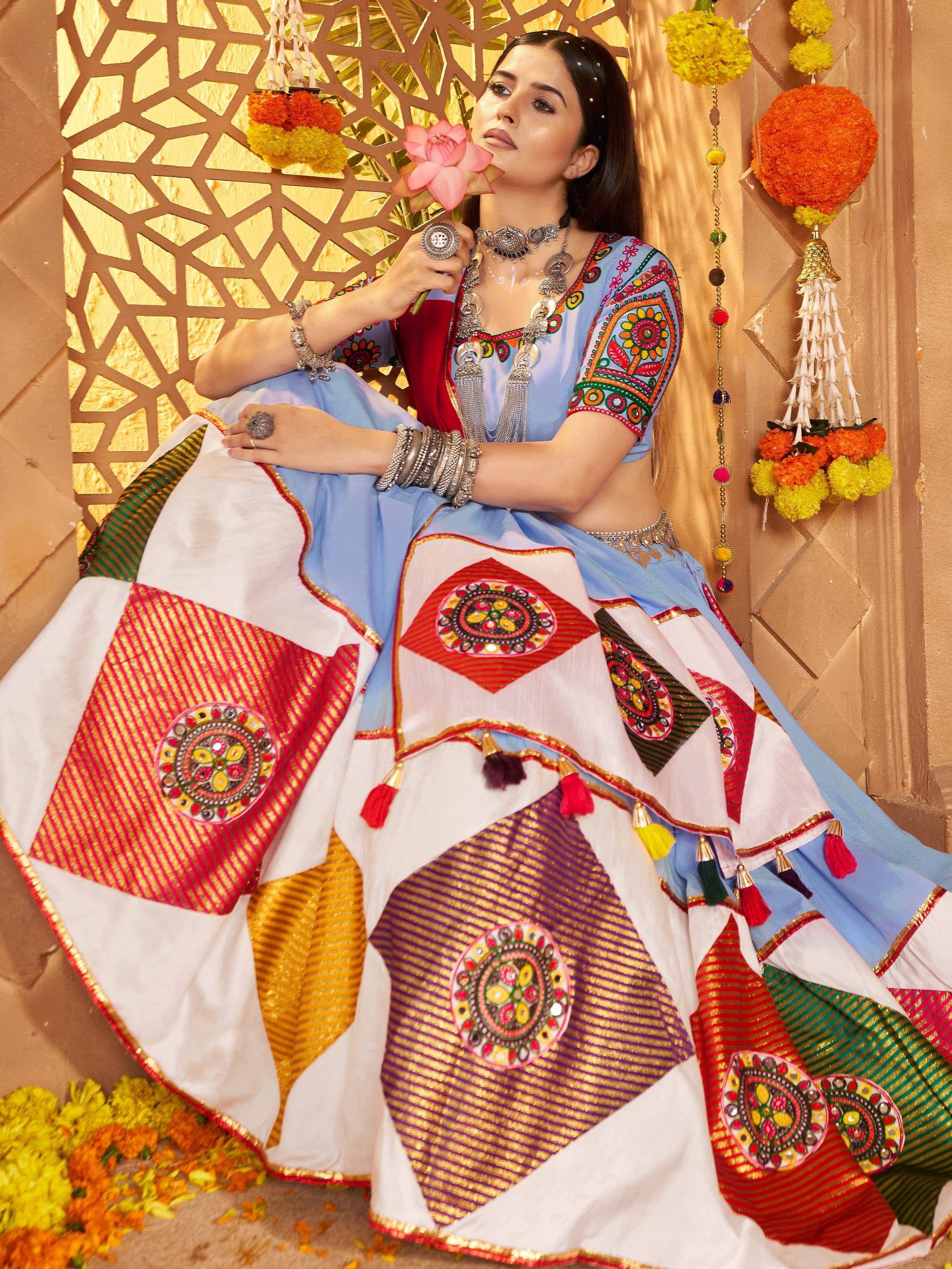 Woman in colorful embroidered Indian lehenga choli with mirror work, holding pink lotus, festive marigold decor