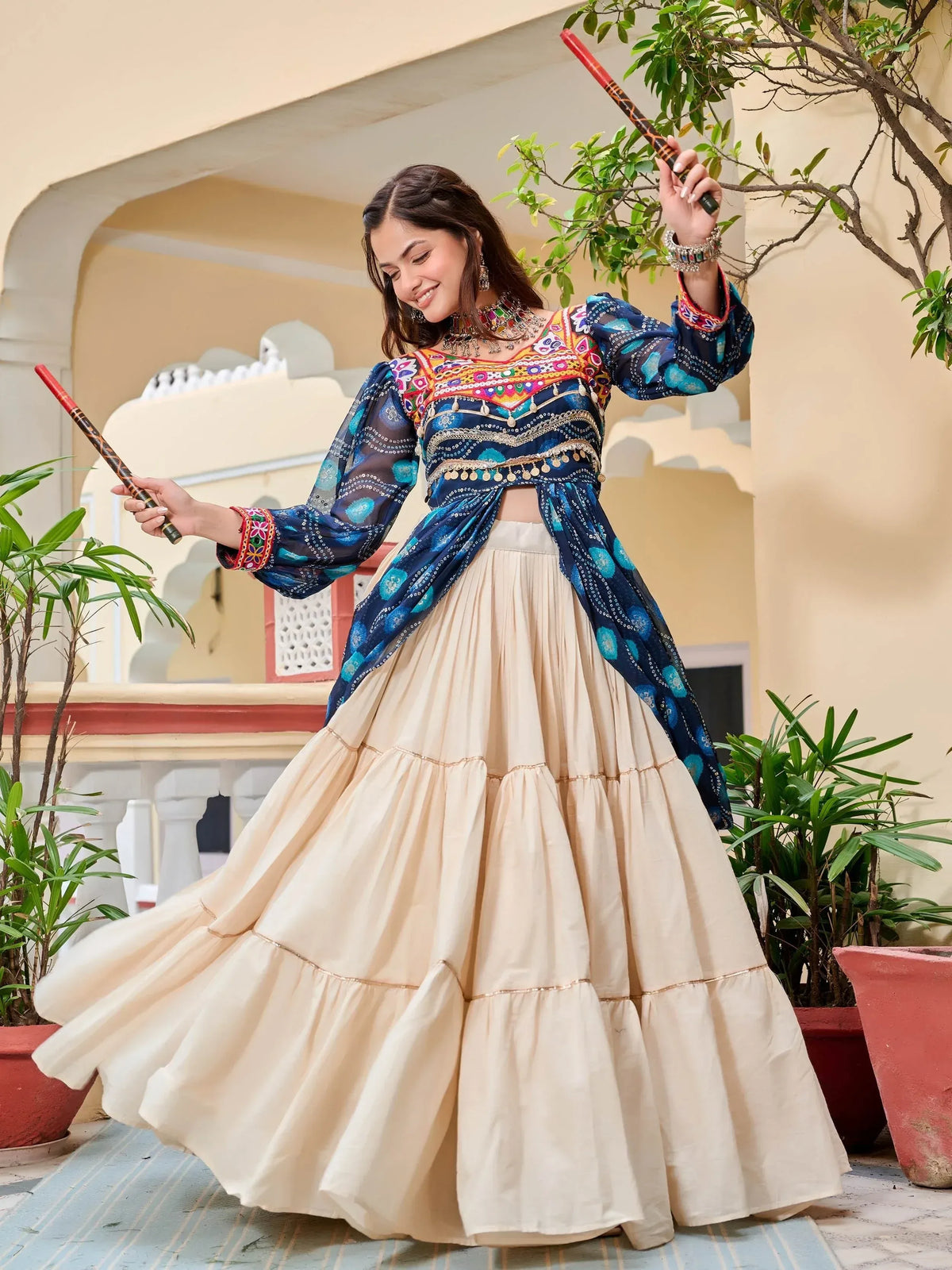 Woman in colorful embroidered blue top and cream tiered skirt, holding dandiya sticks, Indian ethnic wear