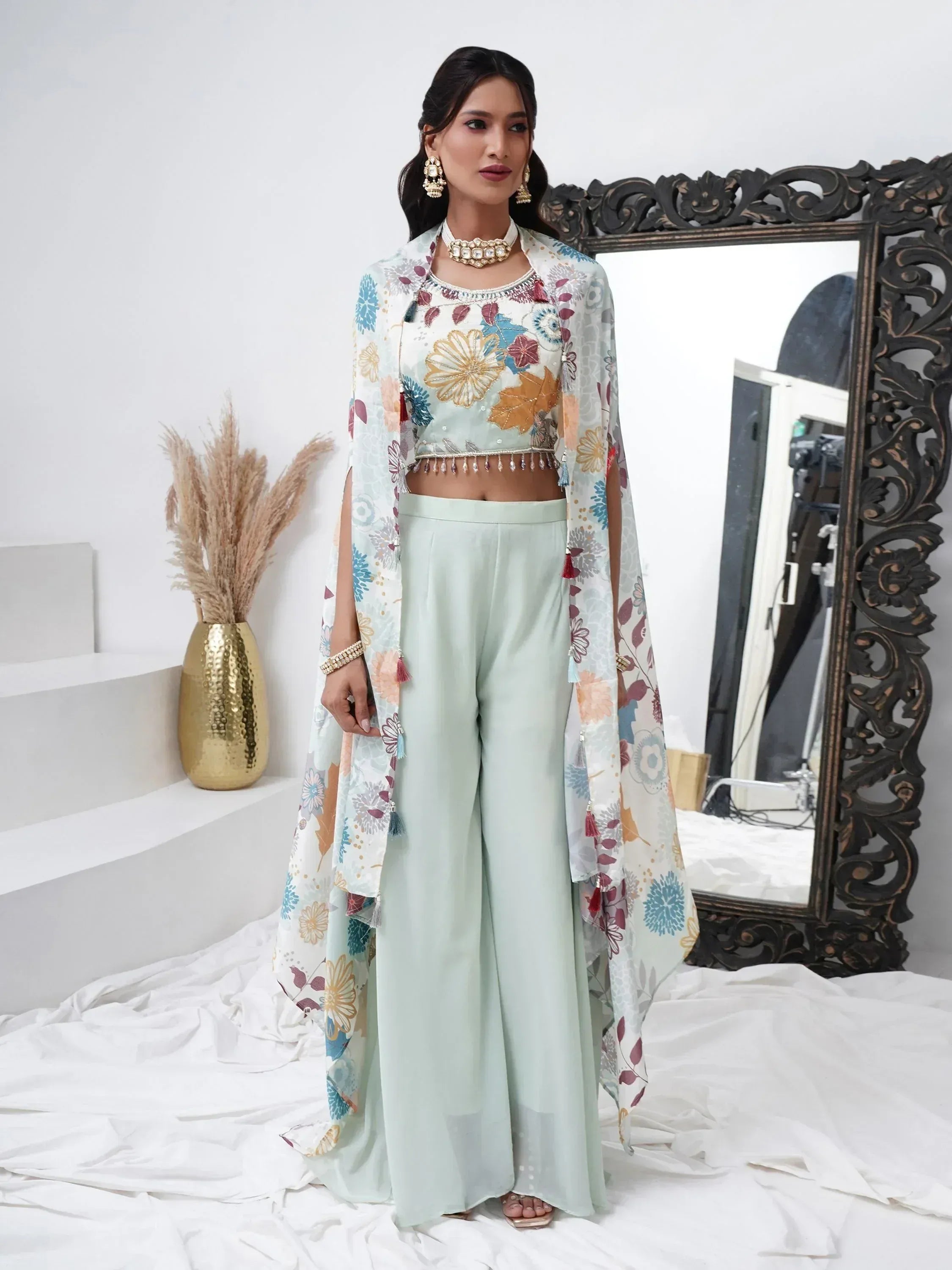 Woman wearing pastel floral ethnic cape set with crop top and palazzo pants, styled with jewelry.