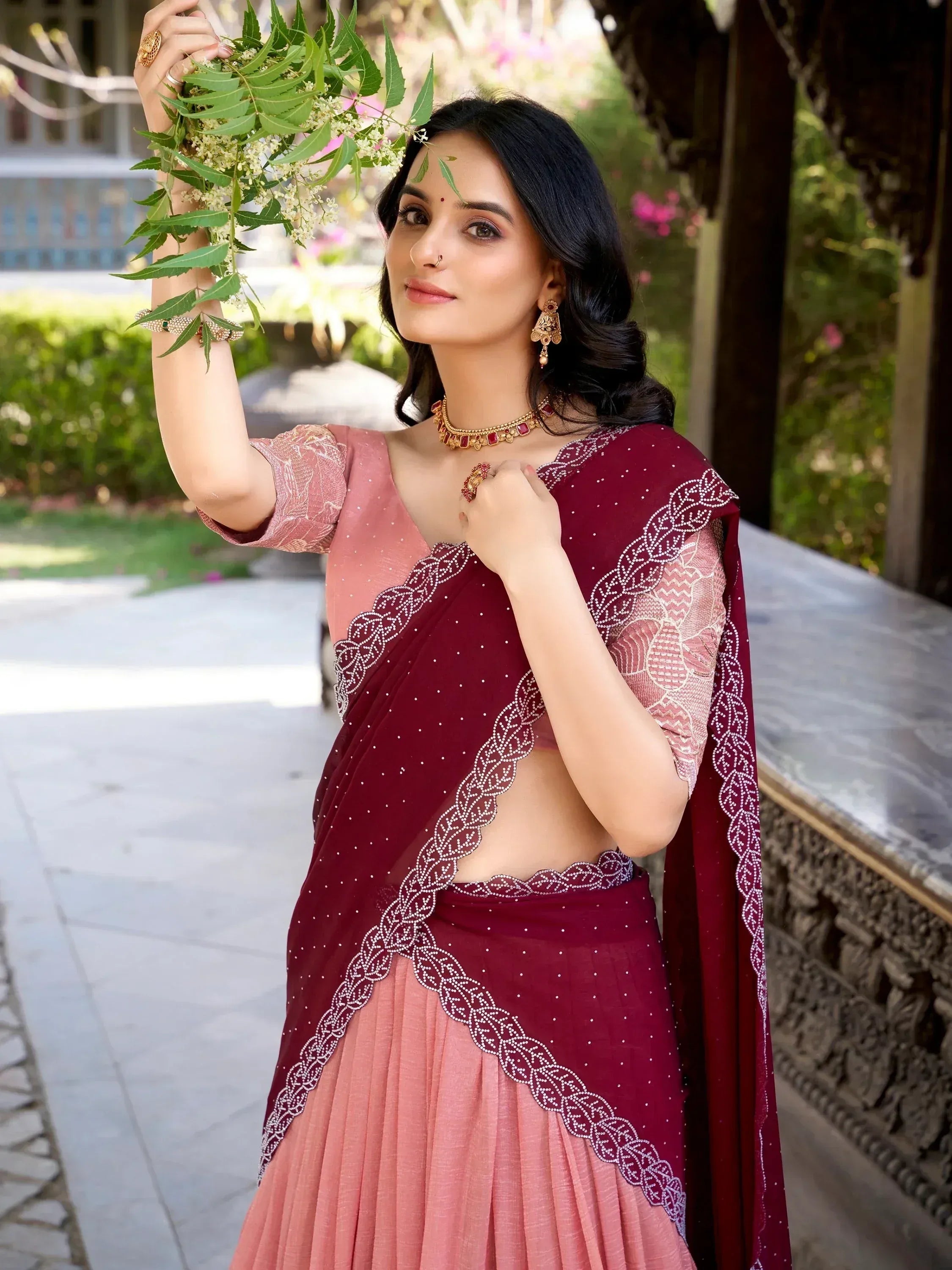 Woman in maroon and pink designer lehenga choli with embroidery, holding green leaves, traditional Indian ethnic wear