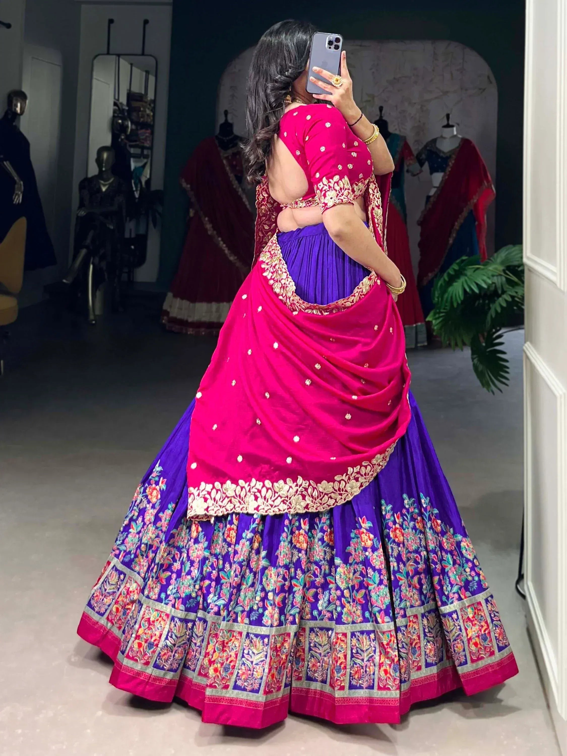 Woman in vibrant pink and purple designer lehenga choli with floral embroidery, taking mirror selfie in boutique