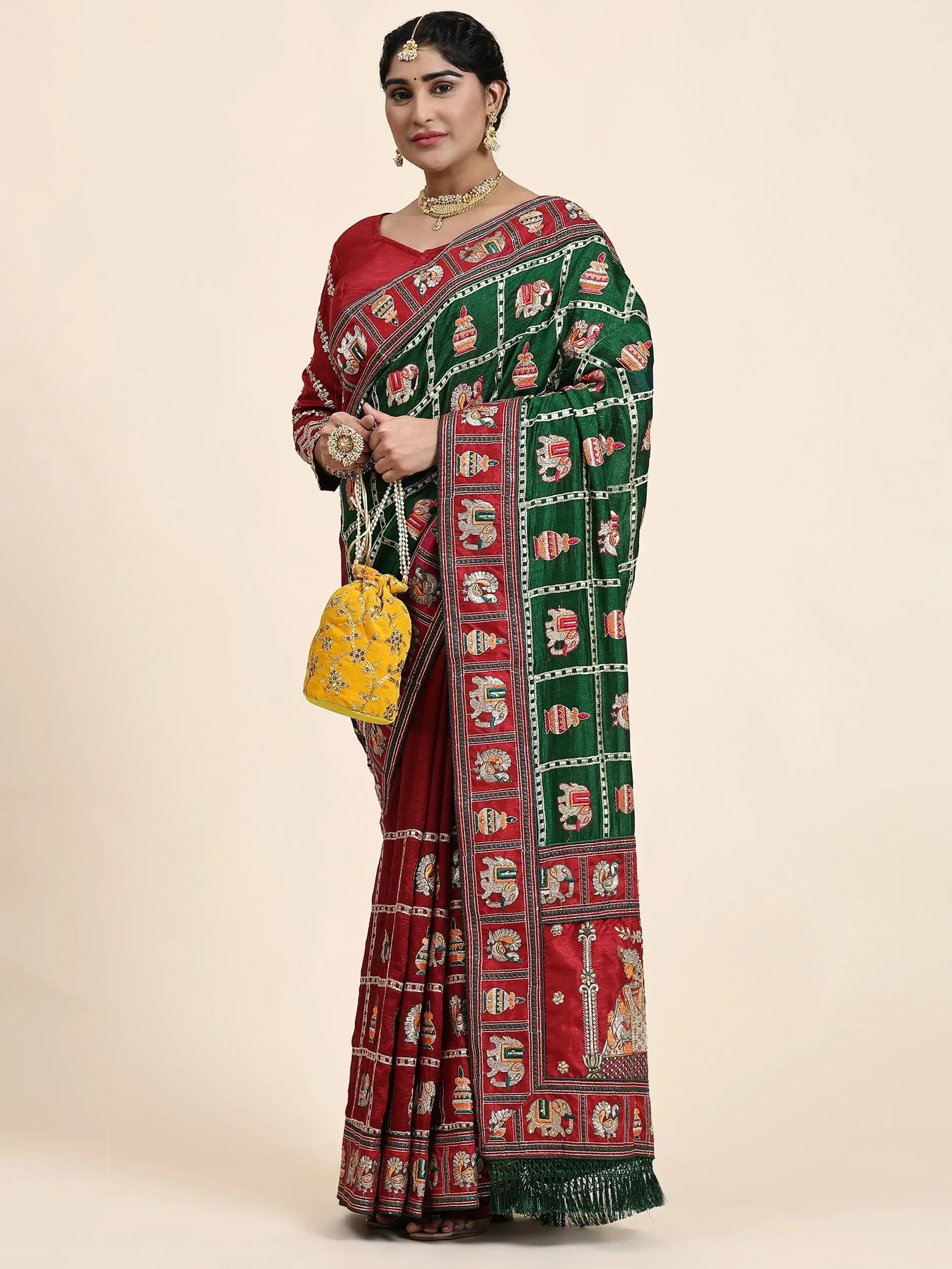 Woman in traditional Indian saree with green and red embroidered elephant and pot motifs, paired with gold jewelry and yellow potli bag.