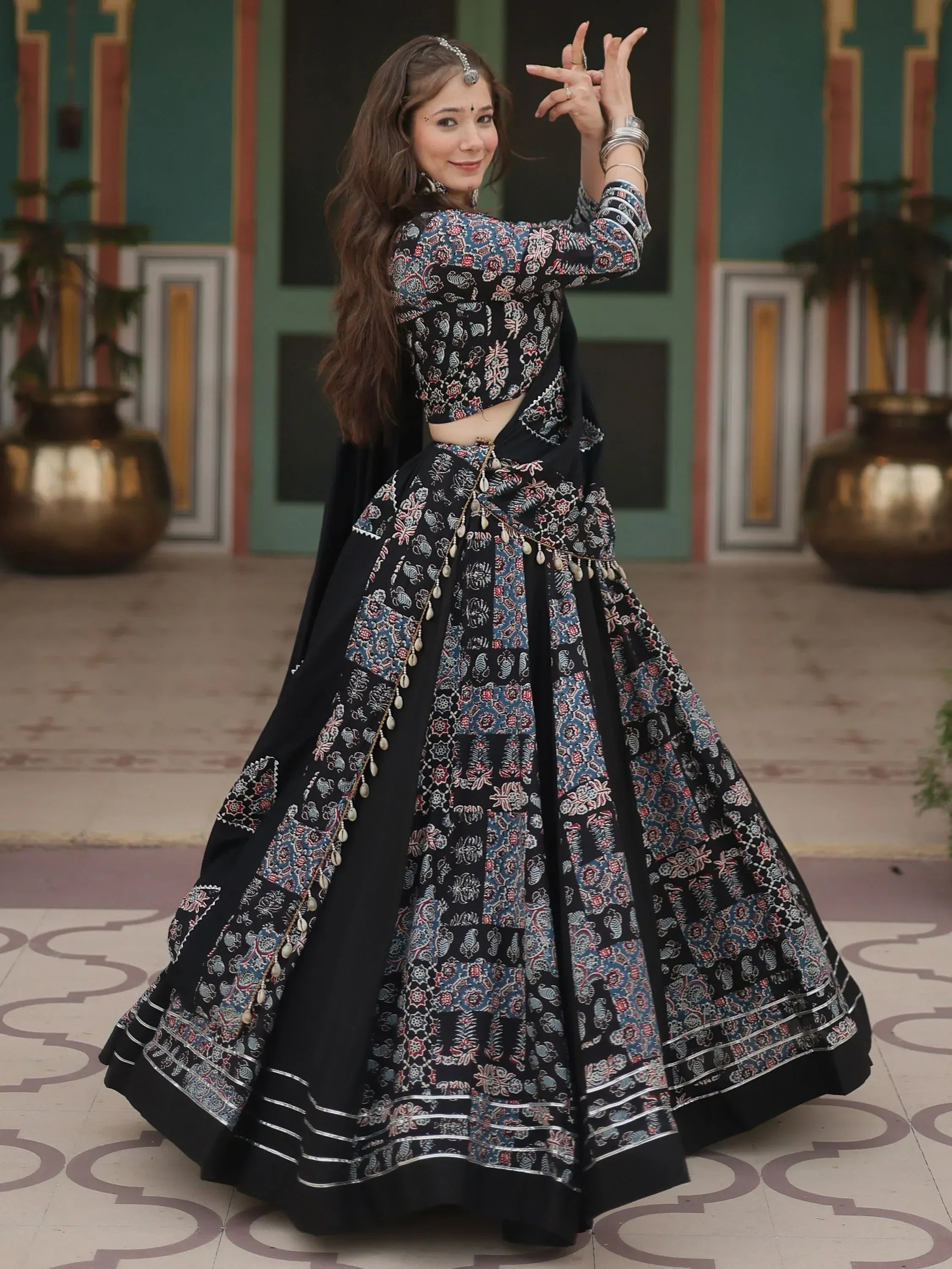Woman in black floral print lehenga choli with mirror work, dancing in traditional Indian attire.