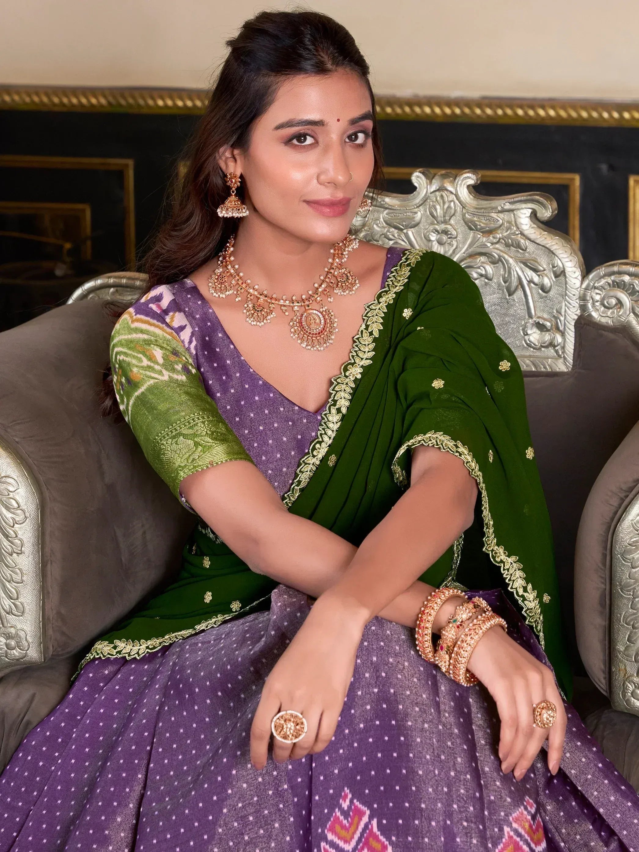 Woman in purple and green ethnic saree with gold jewelry, seated on ornate chair