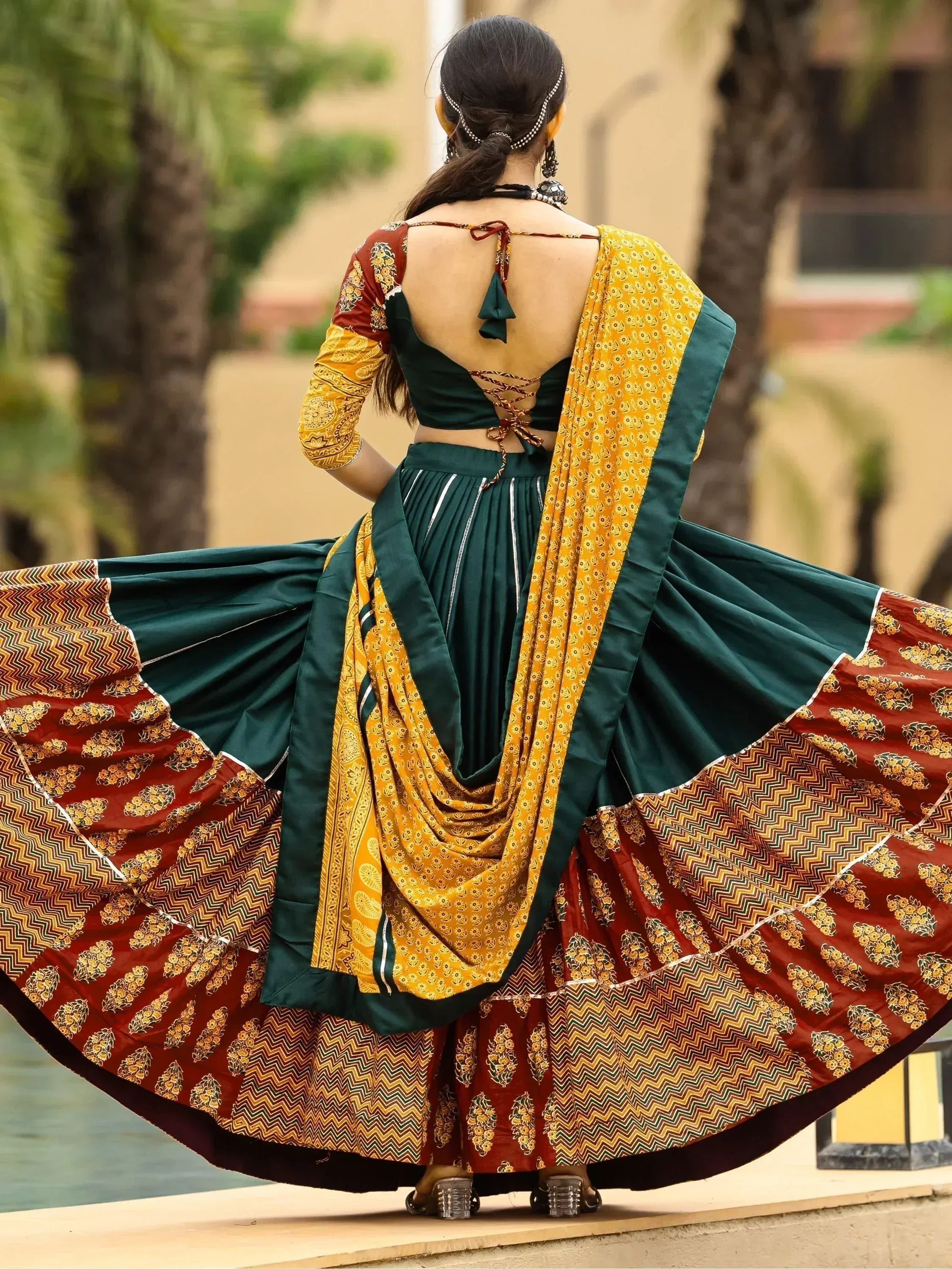 Back view of woman in green and maroon printed lehenga choli with yellow dupatta, twirling outdoors.