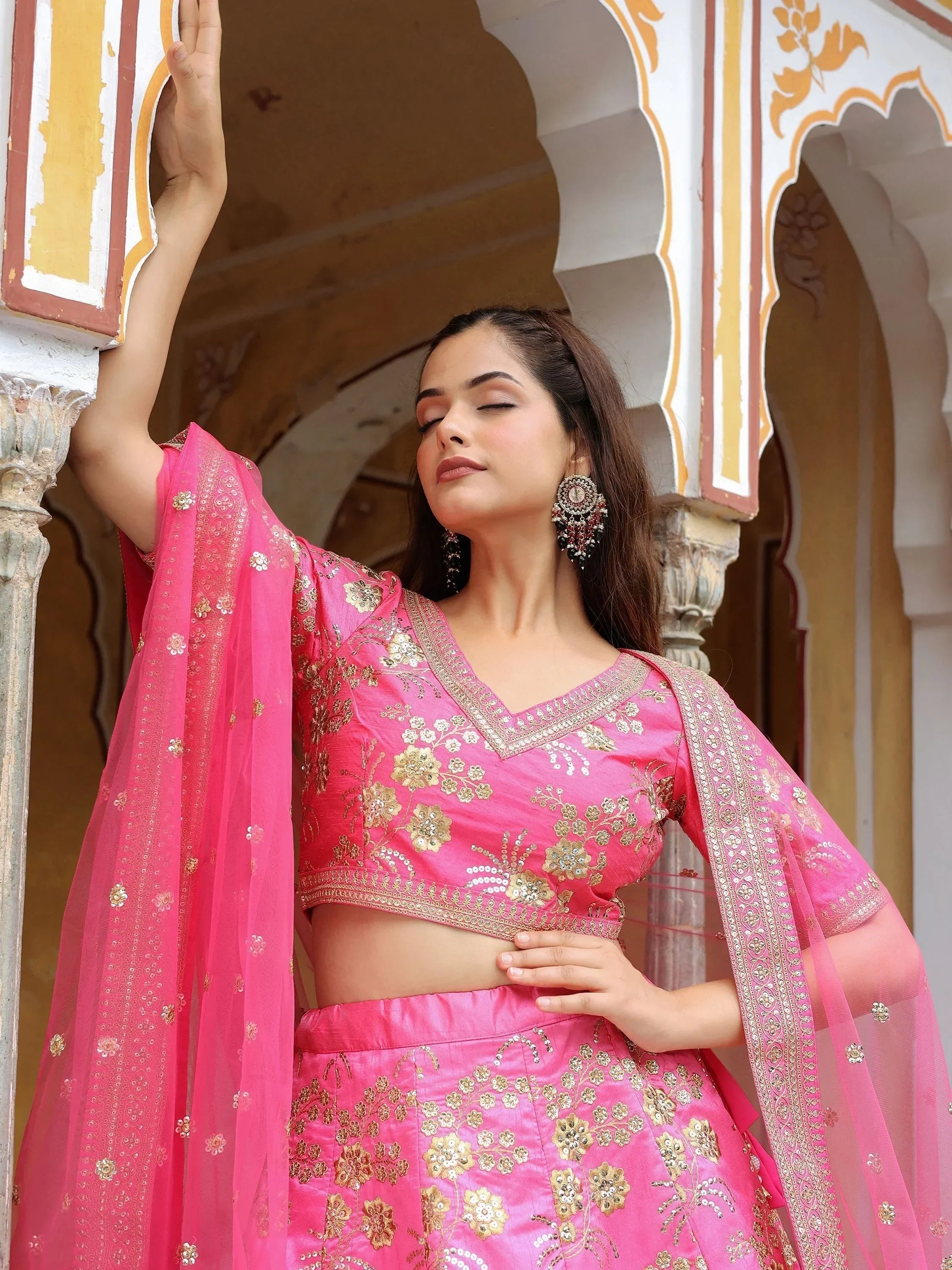 Pink embroidered lehenga choli with gold floral work, sheer dupatta, and traditional Indian architecture backdrop