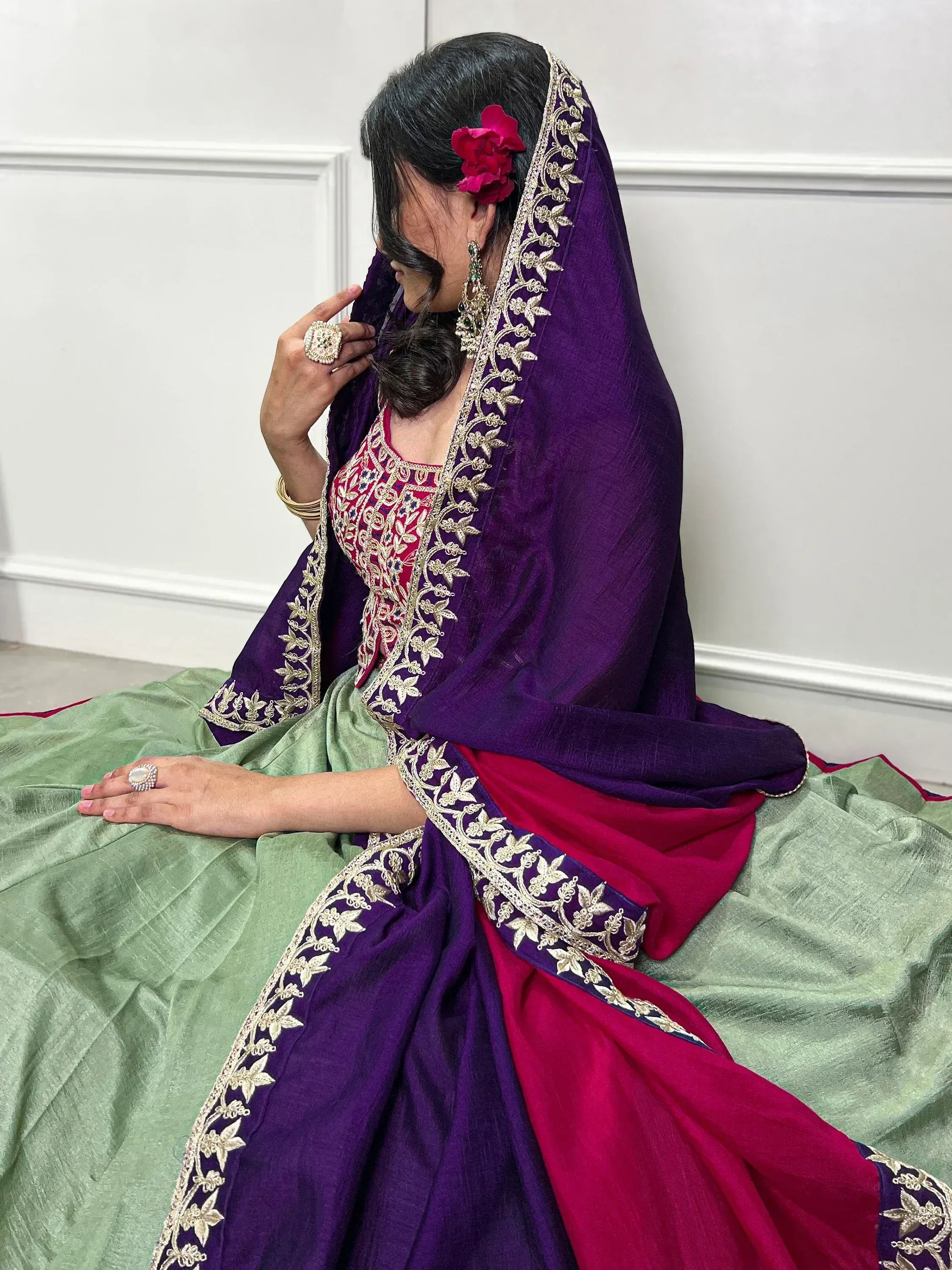 Woman in purple and green designer lehenga choli with gold embroidery, floral hair accessory, and traditional jewelry