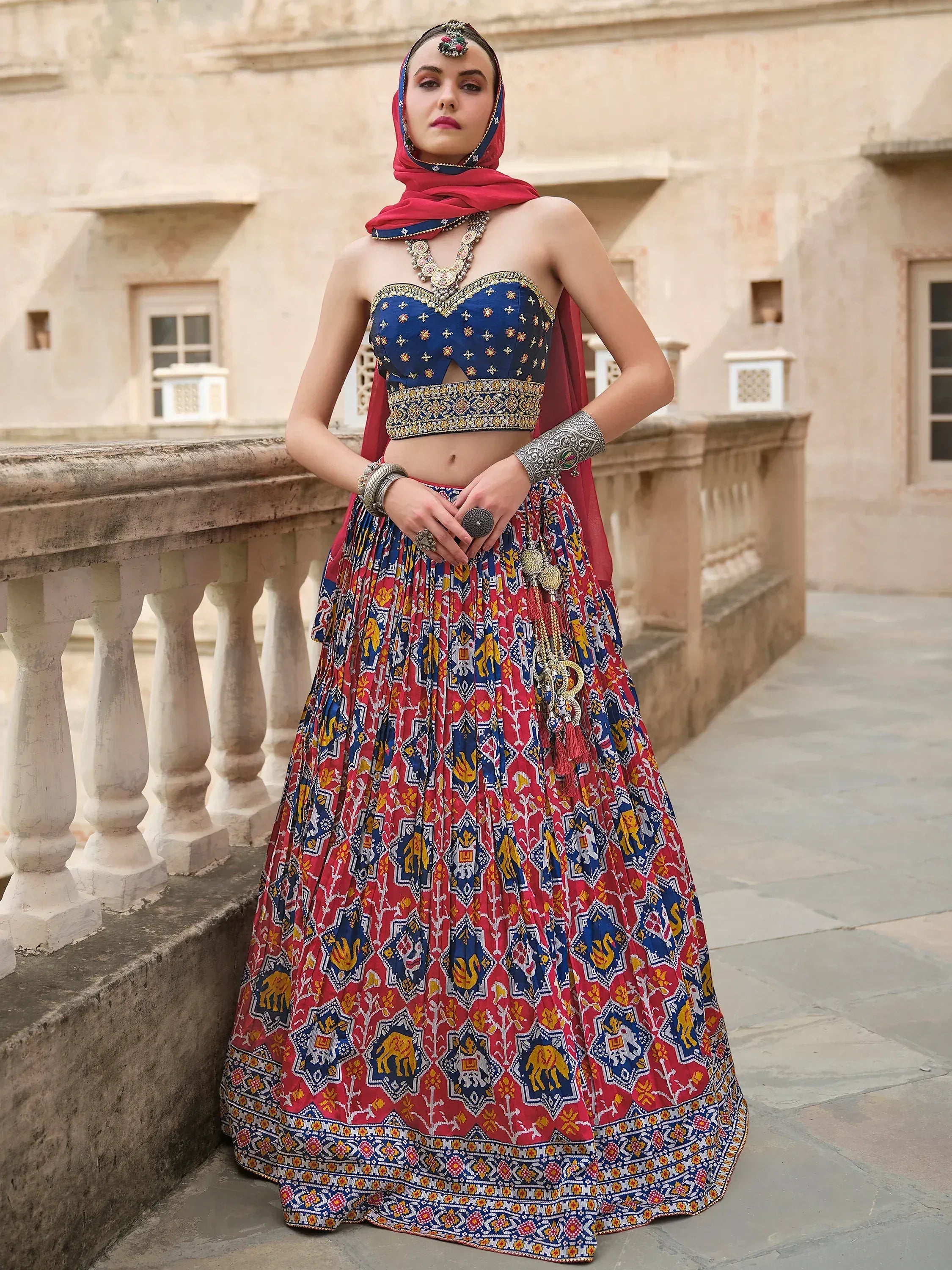 Woman in vibrant red and blue printed lehenga choli with traditional jewelry, standing on a stone balcony