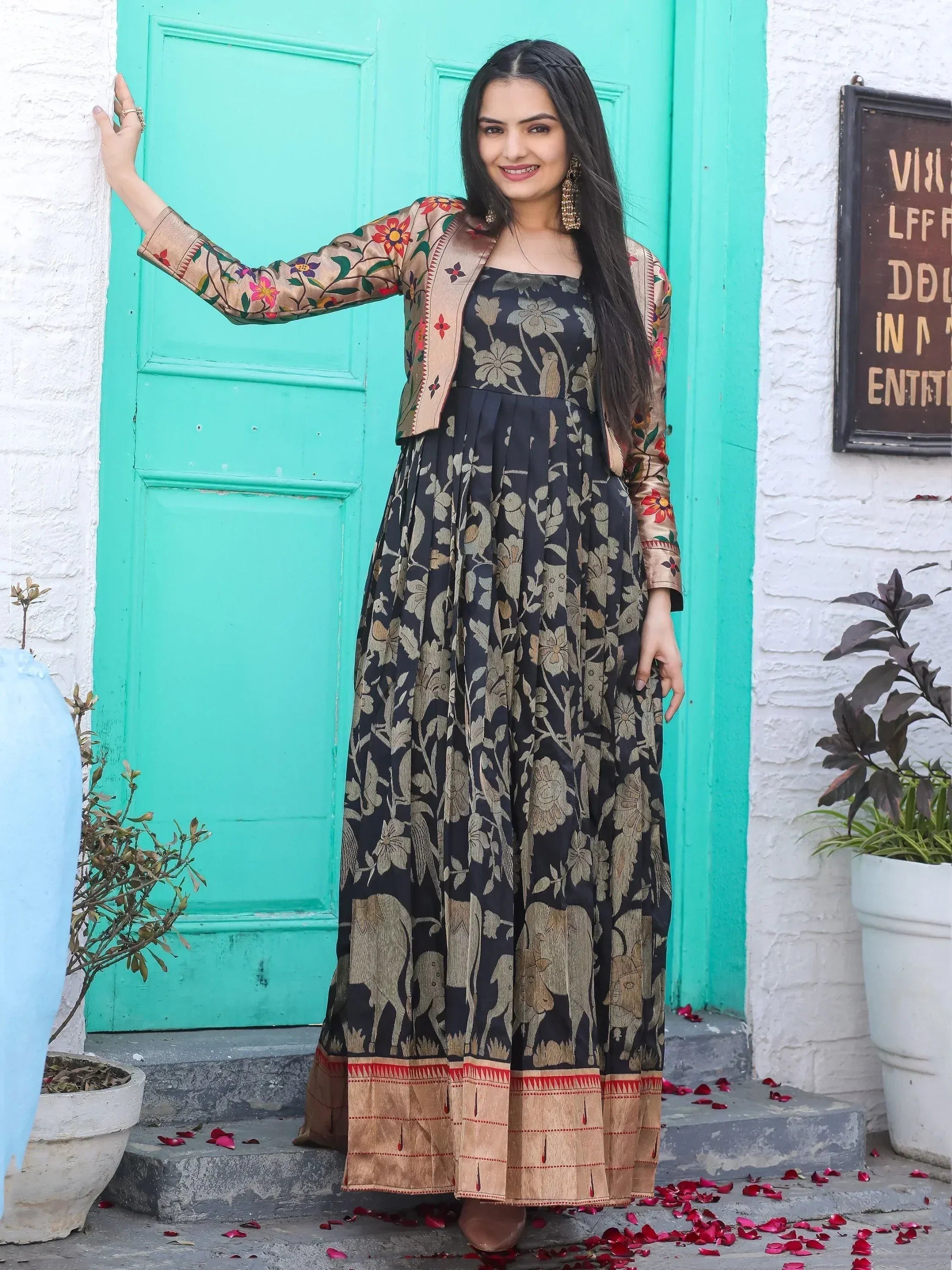 Woman in black floral ethnic gown with embroidered jacket, standing by turquoise door