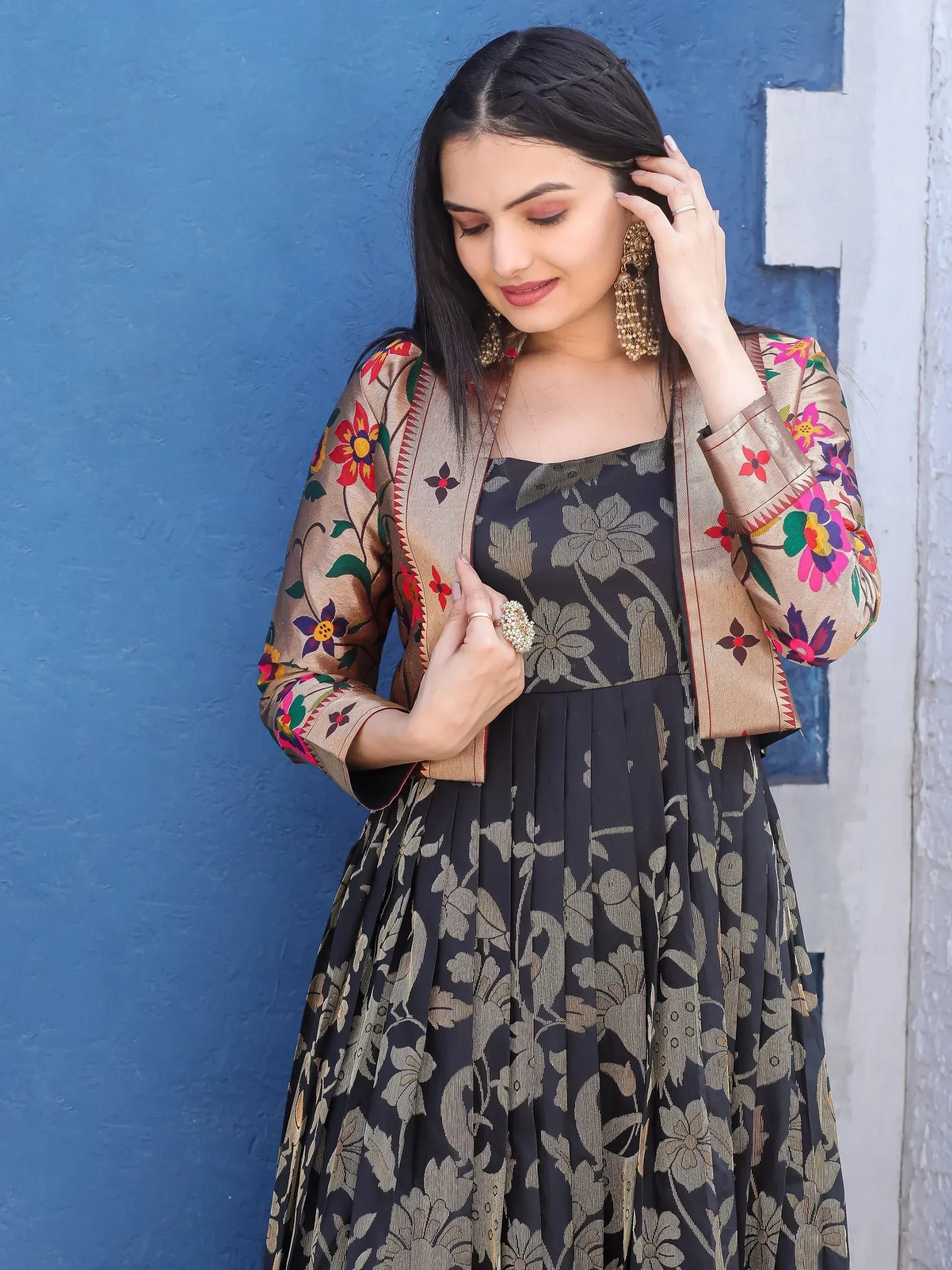 Woman in black floral ethnic gown with embroidered jacket, gold jewelry, against blue wall