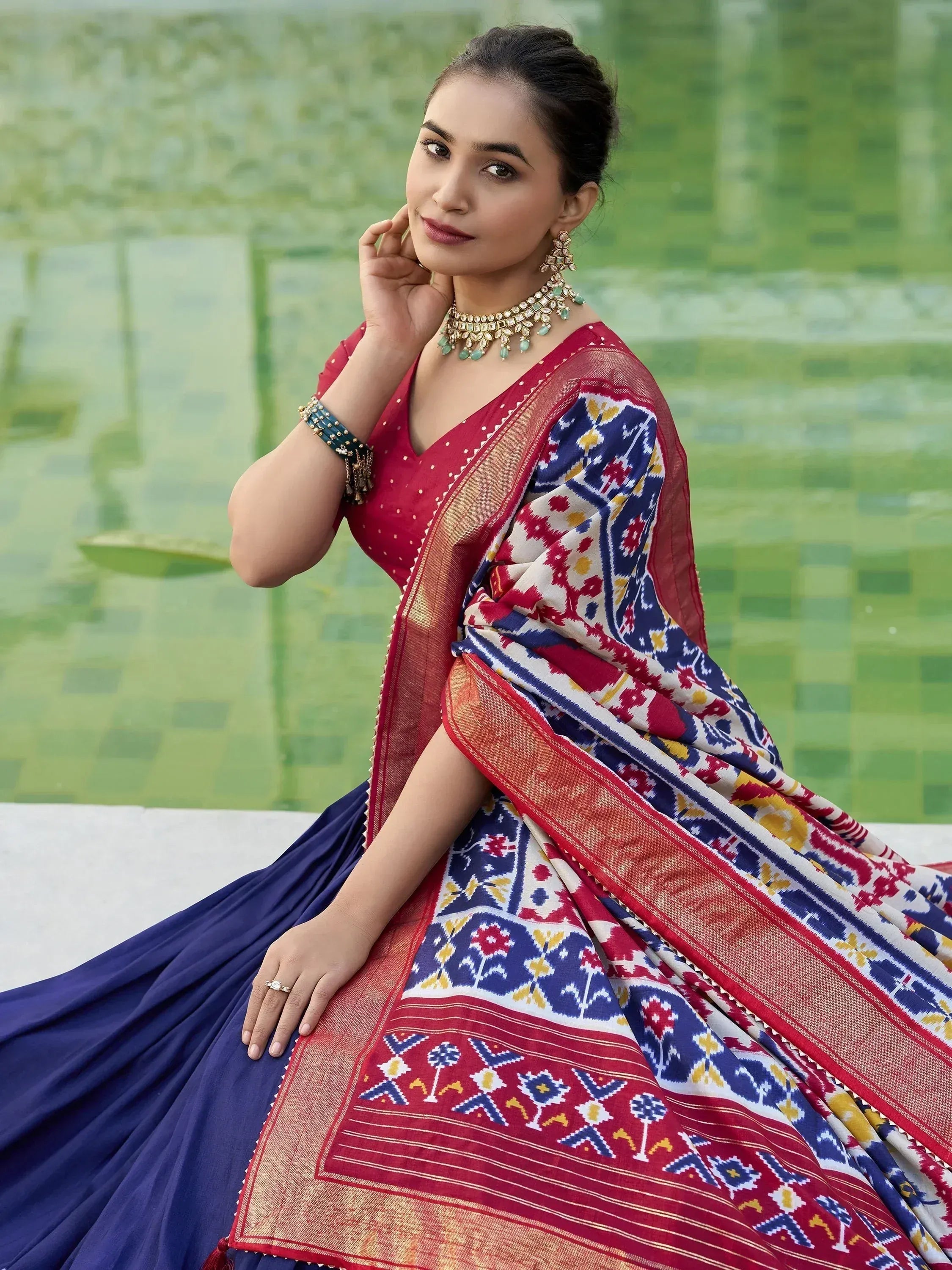 Woman in vibrant red and blue printed saree with gold border, styled with traditional jewelry near pool
