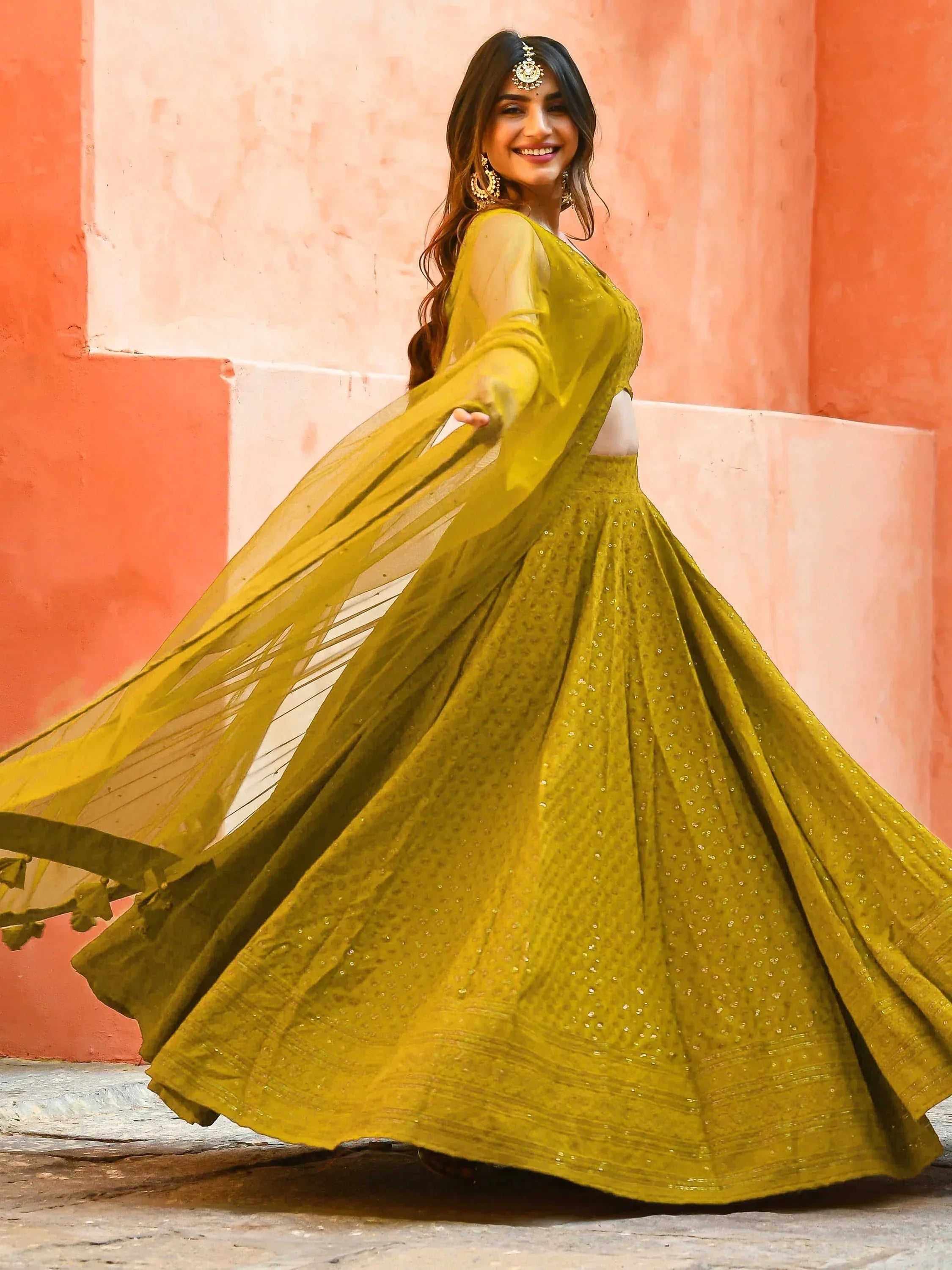 Woman in vibrant green designer lehenga choli with sequins, twirling against pink wall