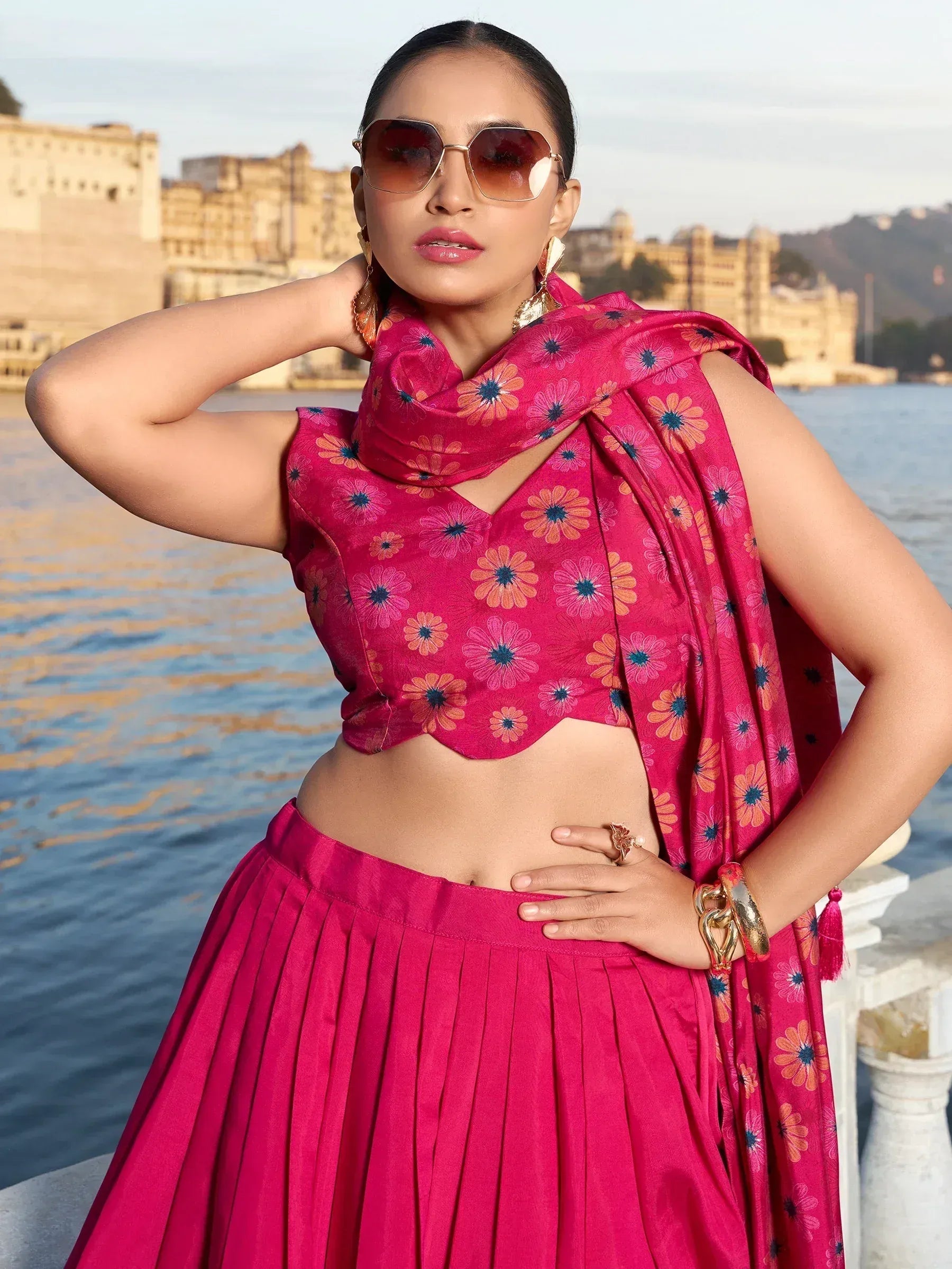 Woman in vibrant pink floral lehenga choli with matching dupatta, styled with sunglasses near water