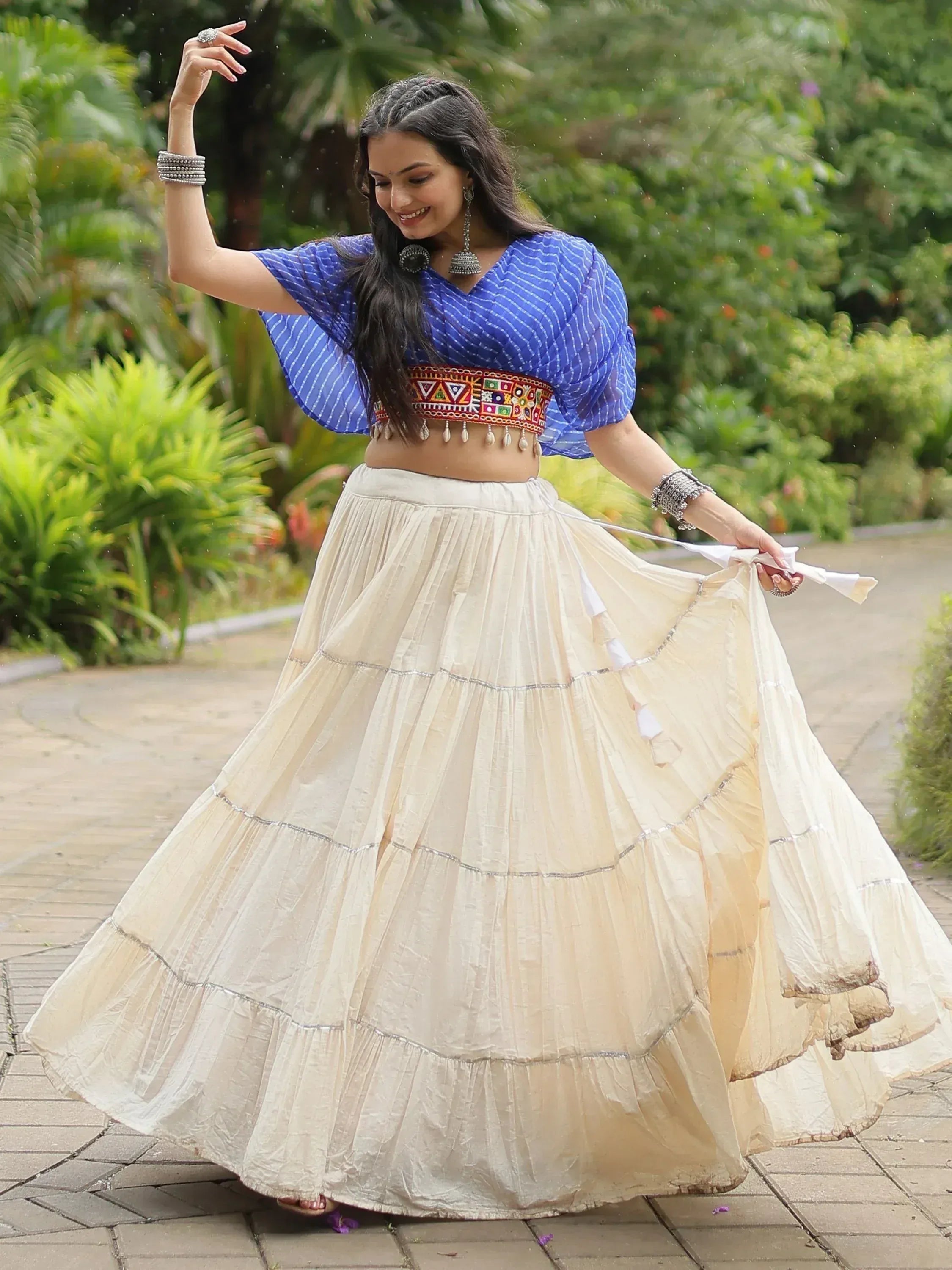 Woman in blue embroidered crop top and cream tiered skirt, dancing outdoors in ethnic Indian wear