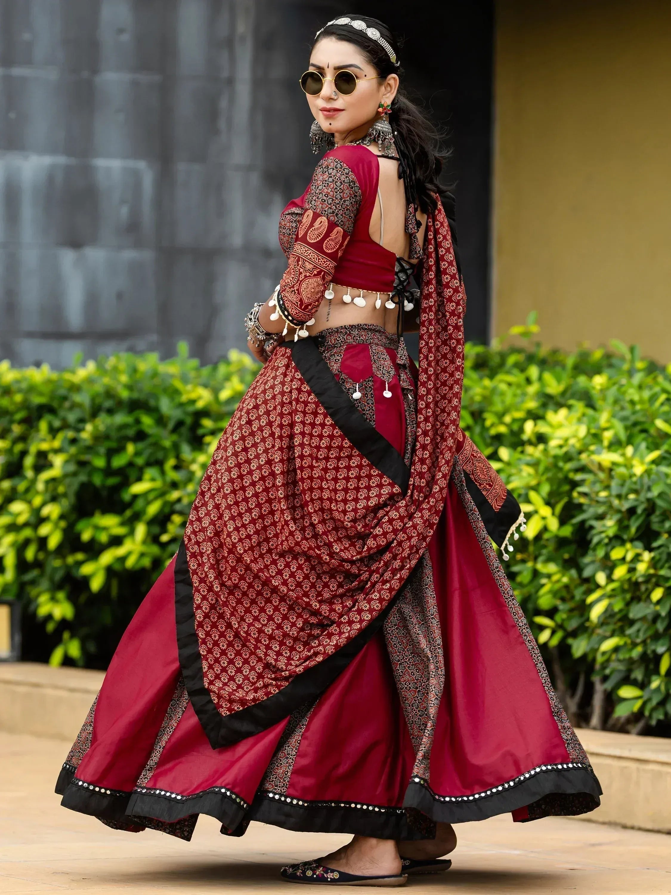 Woman in maroon and black printed lehenga choli with mirror work, ethnic jewelry, and sunglasses